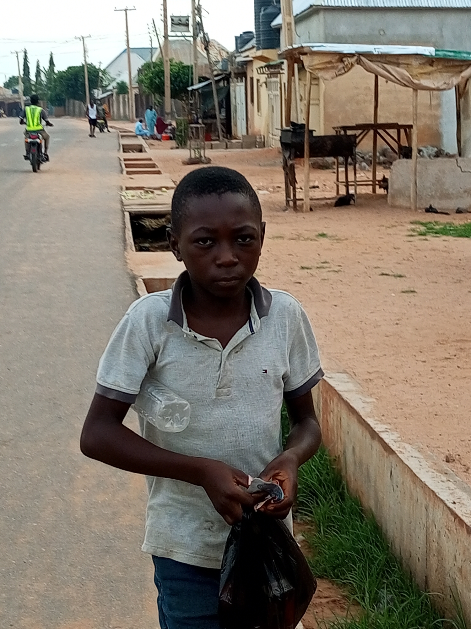 In Gombe, Nigeria, a young boy is captured in a medium shot, standing on a dusty street. He is dressed in a light grey polo shirt with a dark grey collar and sleeve trim, paired with dark blue jeans. In his right hand, he holds a black plastic bag, and in his left hand, he is manipulating a small object, possibly money or another item from the bag. A clear plastic bottle is perched on his right shoulder. The background reveals a street scene with buildings, some appearing to be shops with makeshift awnings, and a person on a motorbike wearing a neon yellow vest in the distance. The ground is a mix of dirt and sand, with a concrete structure and some grass bordering the street. The lighting suggests it is daytime, possibly late afternoon or early morning.
