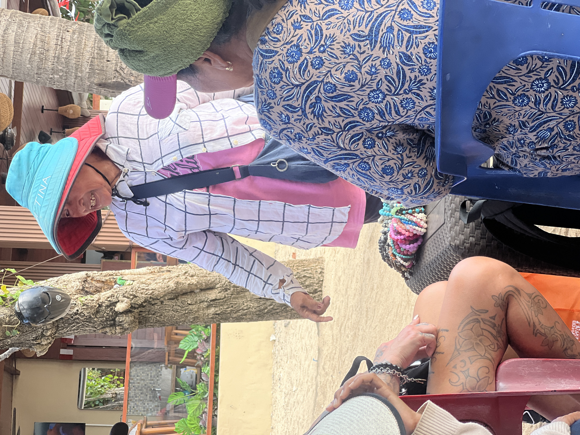 An elderly woman wearing a large blue and red sun hat stands on a sandy beach, her face turned to the left, her mouth open in a smile. She wears a white and black checkered shirt over a pink t-shirt, with a dark blue bag slung over her shoulder. She is standing next to a tree with a black helmet hanging from a branch. To her right, a person with a green towel wrapped around their head is seated, facing away from the camera. In the foreground, a person with tattoos on their legs is seated on a red chair, facing left, their hands resting on their lap. A table with numerous colorful beaded bracelets is situated between the seated individuals. The background shows a building with wooden elements, planters with green foliage, and more sandy beach. The scene appears to be in Kuta, Indonesia, likely a tourist area.