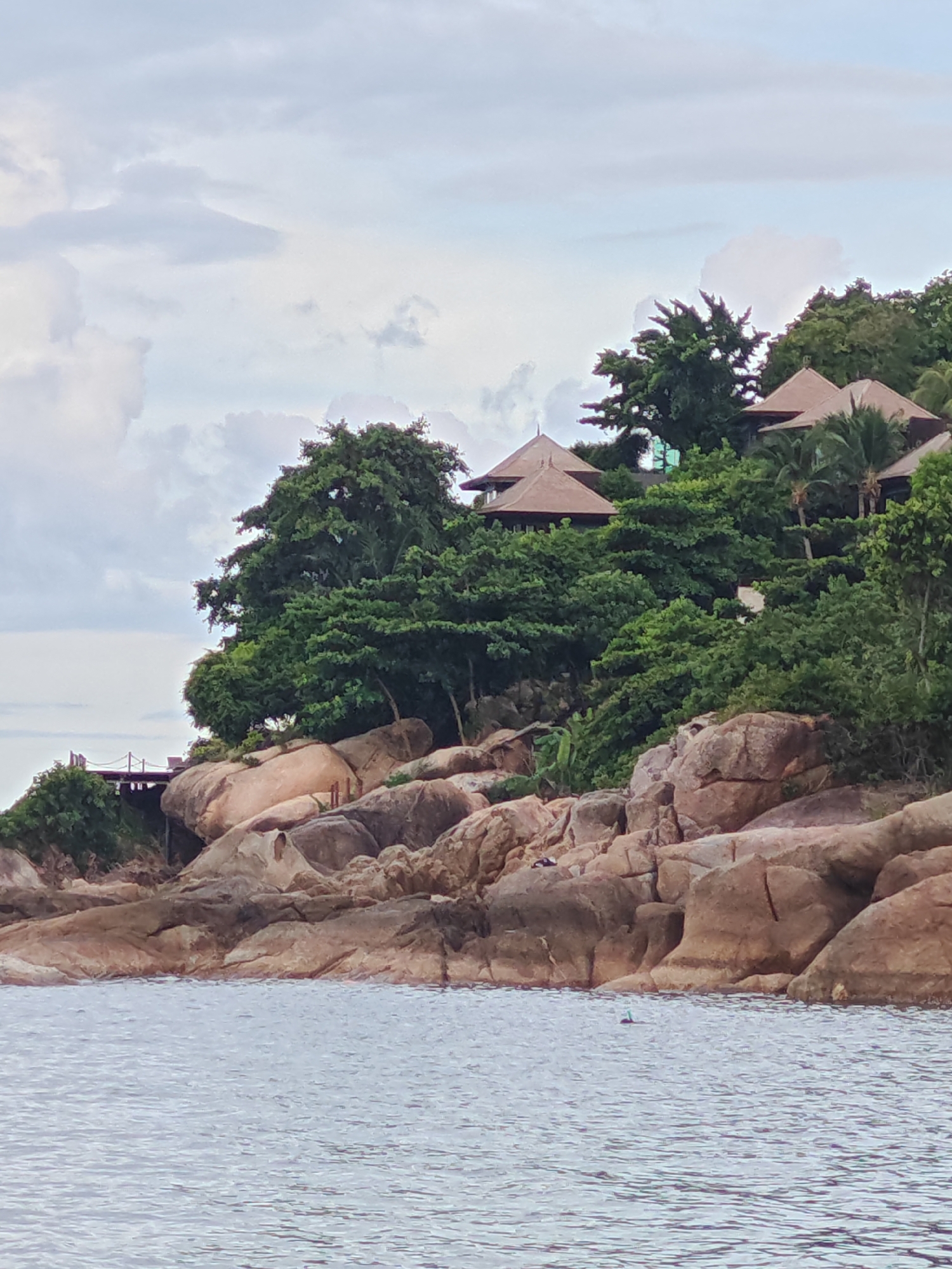 This media file shows a serene coastal scene in Baan Chaweng Noi, Thailand. The foreground is dominated by the calm, rippling surface of the ocean, reflecting the overcast sky. Beyond the water, a rocky shoreline transitions into a lush green hillside. Large, weathered boulders, varying in shades of brown and grey, form the natural embankment.
Perched atop the hillside are several structures with distinctive brown, conical roofs, suggesting resort accommodations or private villas. These buildings are nestled amongst dense, vibrant green trees, creating a harmonious blend of architecture and nature. A wooden walkway or bridge is visible on the left side, leading towards the structures.
The sky is filled with soft, diffused clouds, indicating either early morning or late afternoon, or possibly a generally cloudy day. The weather appears mild, with no harsh sunlight. A lone snorkeler, with a snorkel and mask visible, is in the water, enjoying the ocean. The overall atmosphere is peaceful and tranquil, evoking a sense of relaxation and escape. There is no visible text in the media file.