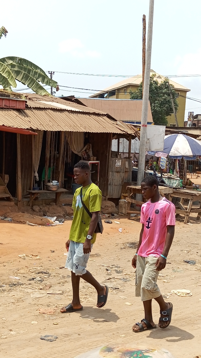 An outdoor daytime scene in Onitsha, Nigeria, depicts a bustling street or market area with two young males walking across an unpaved, sandy dirt ground littered with debris. The background features several informal structures constructed with corrugated metal roofs and wooden supports, suggesting market stalls or small shops. A large banana leaf is visible in the upper left, partially obscuring overhead electrical wires. In the mid-ground, a tall, light-colored utility pole stands prominently. Beyond the immediate structures, a multi-story building with a yellowish-tan roof is visible.
The individual on the left wears a lime green short-sleeved t-shirt with a dark graphic, light blue frayed denim shorts, and black slip-on sandals. He carries a dark shoulder bag and a silver-colored watch on his left wrist, looking forward with a neutral expression. The individual on the right wears a pink short-sleeved t-shirt with a dark emblem (possibly an anchor with a crown), light-colored cargo shorts, and dark two-strap sandals. He has a light blue bracelet on his left wrist and is looking downwards.
Behind the individuals, several wooden tables and a blue and white striped market umbrella indicate vendor activity. One corrugated metal structure has hanging fabric strips as a makeshift curtain and a metal bowl on a wooden bench. The ground is uneven and shows signs of wear and scattered refuse, including plastic and other discarded materials. The two males are walking parallel to each other, not directly interacting, moving from the left side of the frame towards the right.