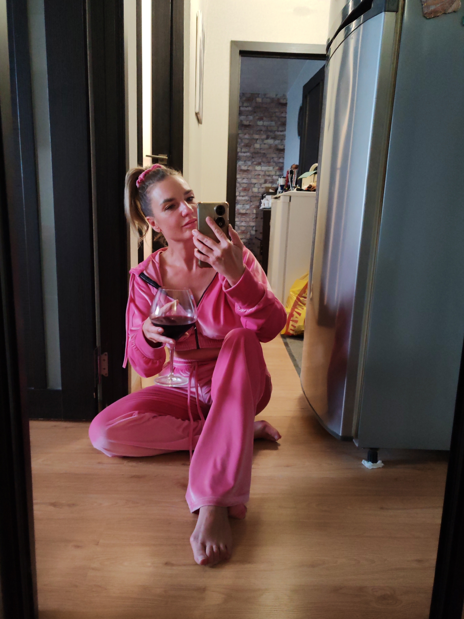 A woman is taking a selfie with her phone while sitting cross-legged on a wooden floor. She is holding a glass of red wine in her left hand. The woman is wearing a pink sweatsuit and has her hair pulled back in a ponytail with a pink scrunchie. Behind her is a refrigerator and two doors. One door is partially open, revealing a room with brick walls and a white cabinet. The scene appears to be indoors, possibly in an apartment or home. There is no specific evidence to support that the location is Petropavlovsk-Kamchatsky, Russia.