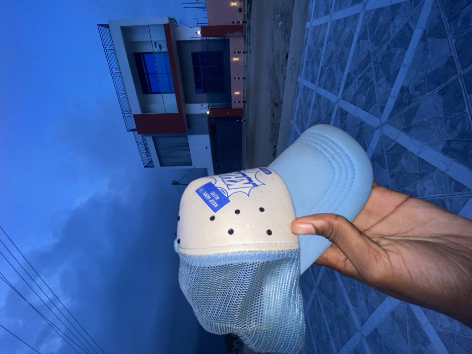 A hand holds a light blue and beige trucker hat against a backdrop of a modern two-story building. The hat has a mesh front and a blue patch with the words "Keep Hope Alive". The building features white walls with red accents and blue reflective windows. The ground has a patterned tile surface. The sky is a dark blue, suggesting either dusk or dawn. Power lines are visible against the sky on the left side of the frame.