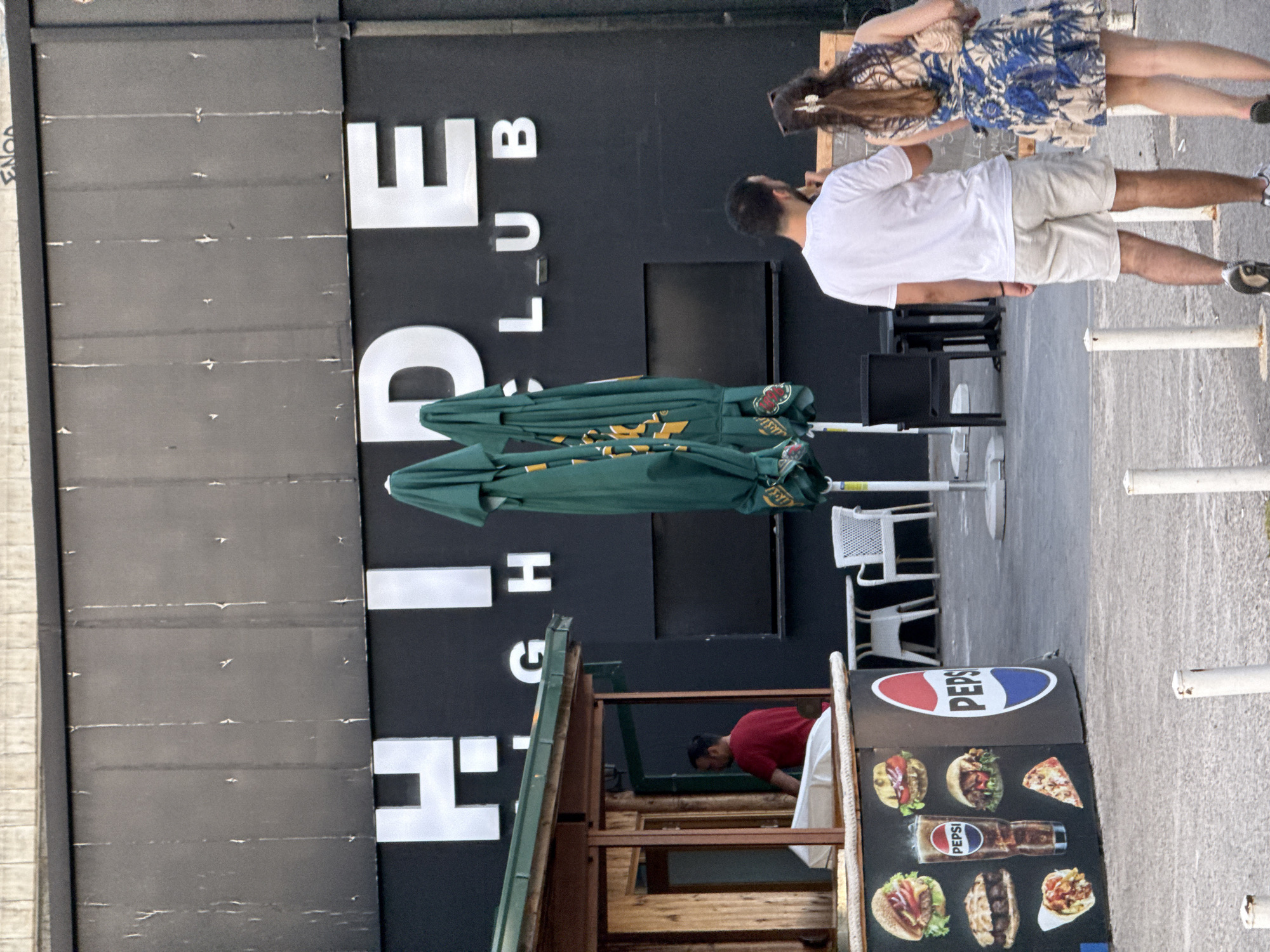 In Budva, Montenegro, a building with the illuminated sign "HIDE CLUB" is visible. In the foreground, there is a food stand with images of burgers, a glass of Pepsi, and a slice of pizza. Two people, a man and a woman, are walking away from the camera towards the right side of the frame. Several green umbrellas are stacked to the left of the building. In the background, there are white tables and chairs. The overall environment suggests an outdoor area of a venue or establishment.
