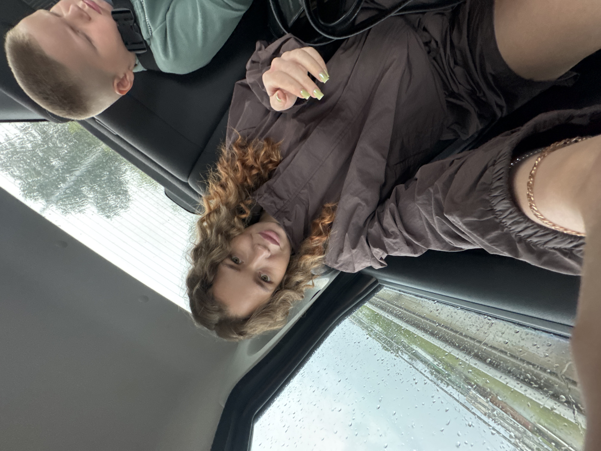 In the rear passenger seats of a vehicle, a young woman with curly brown hair and light-colored nail polish is taking a selfie. She is wearing a brown jacket and dark shorts. To her right, a young boy with a buzz cut is asleep in a car seat, wearing a light green jacket. Rain is visible on the car window to the woman's left, obscuring the view of the outside environment, which appears to be a grassy area with some trees. The interior of the car is dark, with black seats. The location is identified as Chaikovsky, Russia.