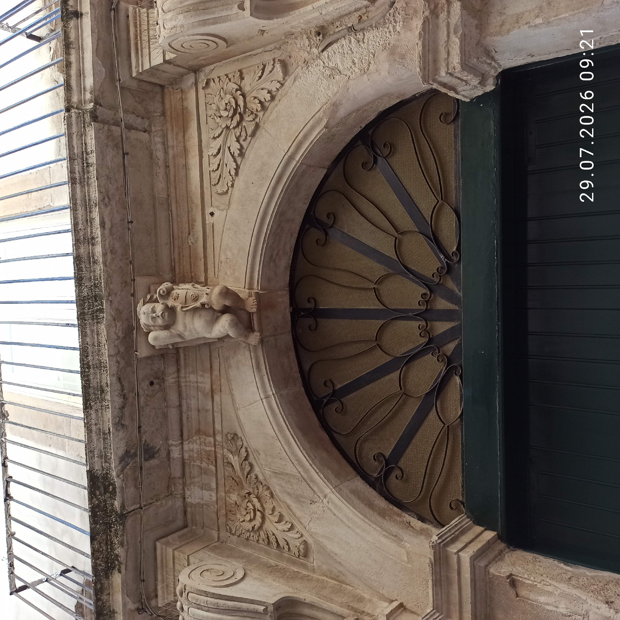 The image shows a close-up, low-angle view of the ornate entrance to a building in Bitonto, Italy. The entrance features a large, dark green wooden door surmounted by an arched transom window with decorative wrought-iron grilles. Above the transom, the building's facade is adorned with stone carvings. At the apex of the arch, a small stone cherub or putto is depicted holding a shield. Flanking the cherub are intricately carved floral motifs on either side of the arch. The stonework appears aged and weathered. A balcony with metal railings is visible at the top of the frame. The timestamp "29.07.2026 09:21" is superimposed on the lower right portion of the image.