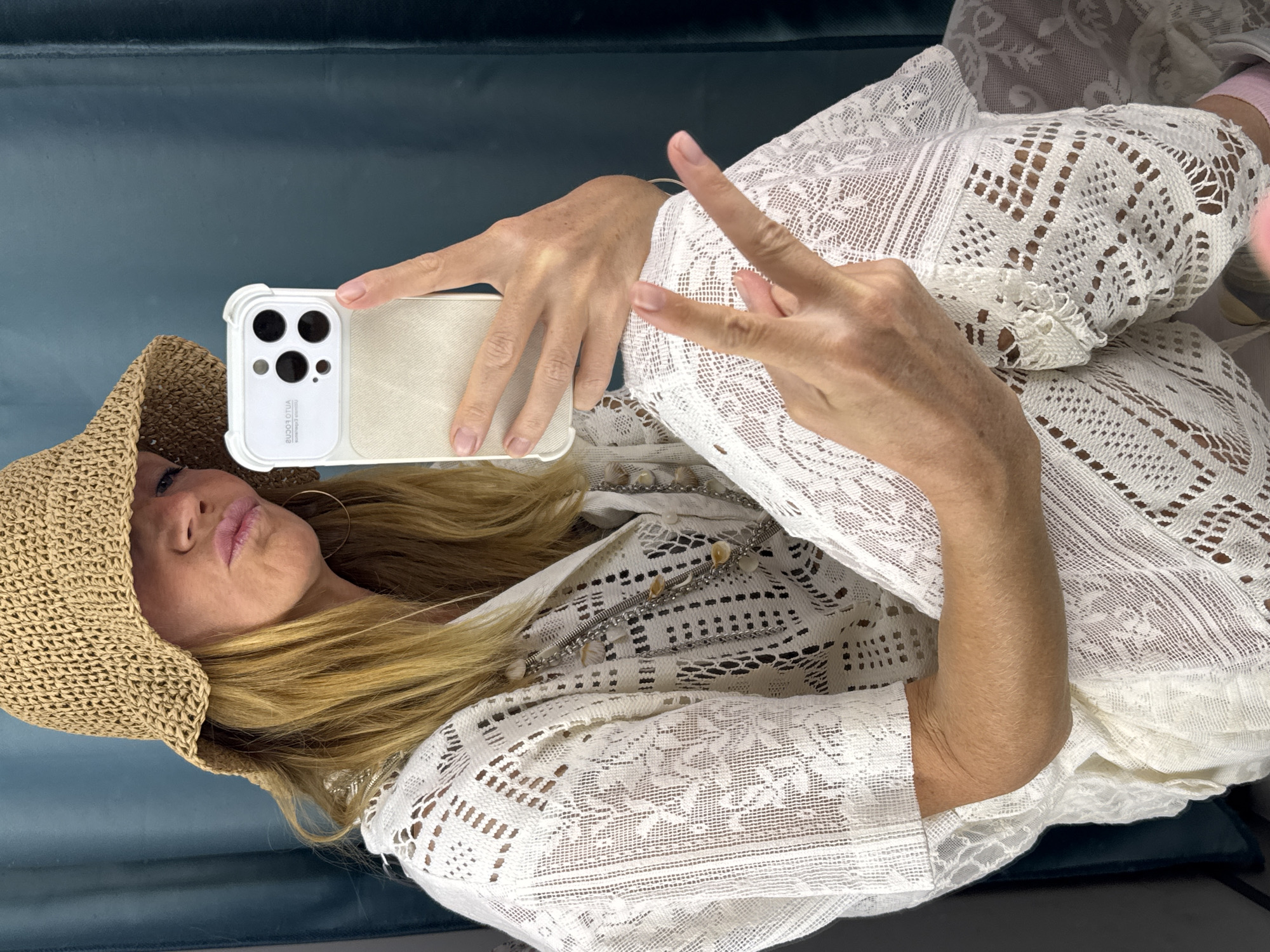 A woman wearing a woven hat and a white lace outfit is holding a cell phone and making a peace sign. The background appears to be a blue-green surface. The location in the city of Saint Petersburg, Russia, cannot be determined from the image.