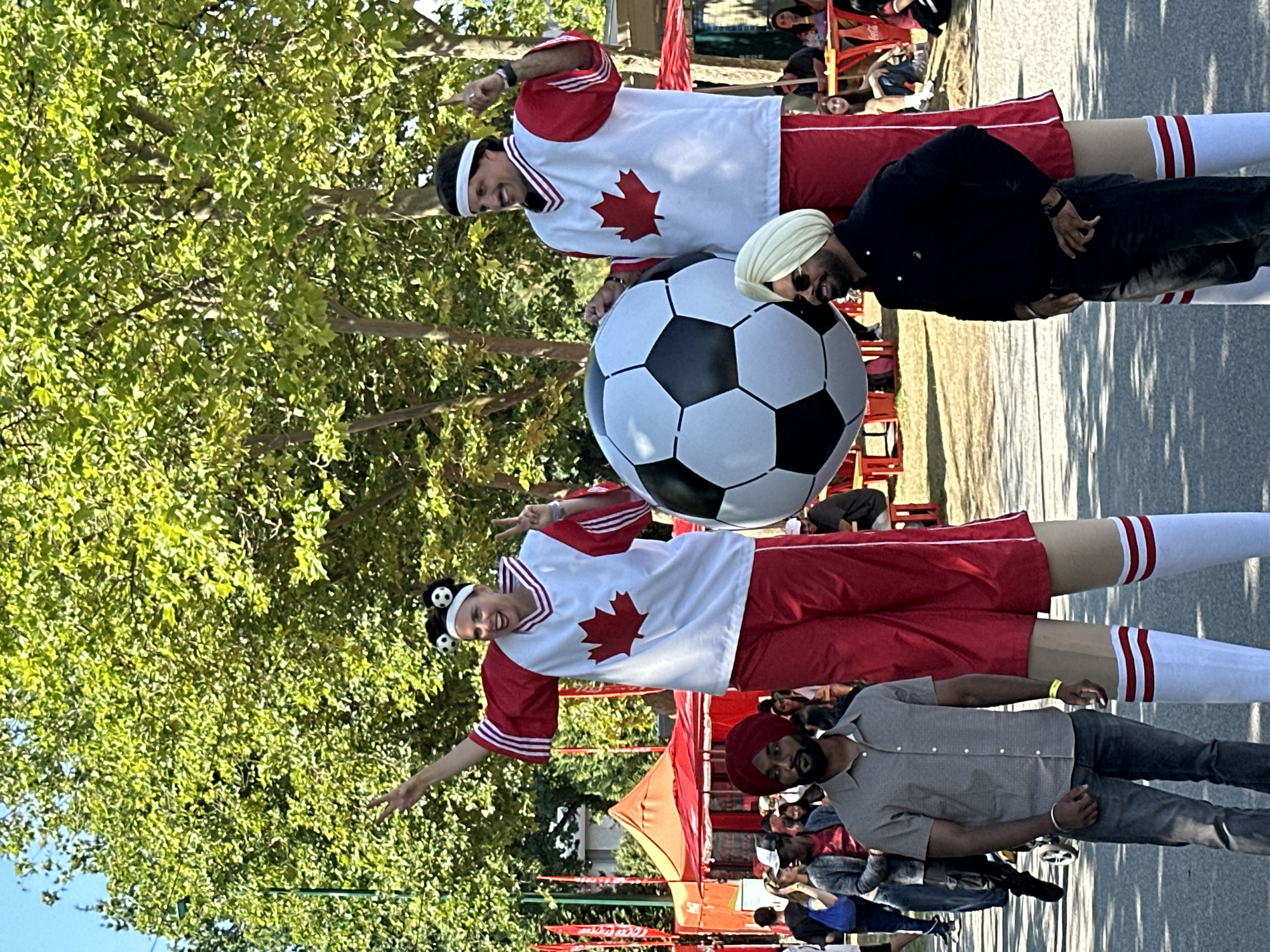 In Vancouver, Canada, on a sunny day, two performers on stilts, dressed in Canadian-themed red and white athletic attire featuring maple leaves, pose with a giant inflatable soccer ball. The female performer on the left wears a headband with small soccer ball accents and makes a peace sign with her hand. The male performer on the right also gestures upwards with one hand. Both wear white headbands. In the foreground, two men in casual wear stand. The man on the left wears a grey button-down shirt and jeans, while the man on the right wears a black shirt and turban. The background features trees, tents, and other attendees at an outdoor event.