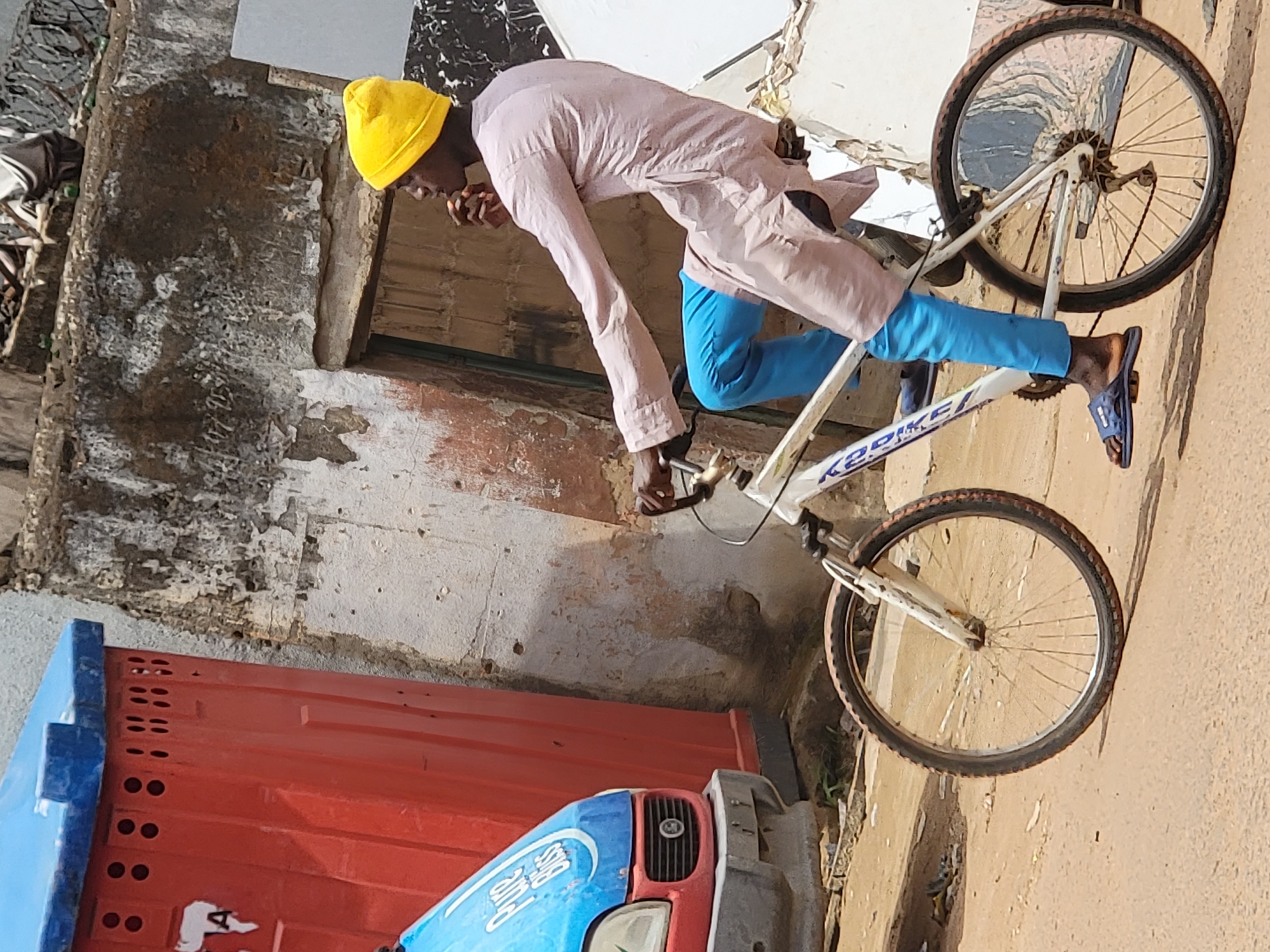 A street scene in Maiburiji, Nigeria, unfolds under bright daylight. A dark-skinned adult male, wearing a yellow beanie, a light beige or pinkish-brown long-sleeved tunic, bright blue pants, and blue open-toed sandals, is seated on a white "KED BIKE" bicycle. His right hand is near his mouth, and he looks to the right. The bicycle is stationary or moving slowly, with its front wheel visible on the unpaved ground.
To the left of the cyclist, the front portion of a light-blue vehicle, likely a van, is visible, featuring "Pure Bliss" branding on its bonnet and a red grille. Directly behind the vehicle and to the left of the cyclist, stands a tall, red, rectangular structure with a blue top, appearing to be a portable utility unit or public restroom, marked with a white "A" and multiple ventilation holes.
The background consists of a heavily weathered wall, possibly concrete or stucco, displaying significant peeling paint, discoloration, and structural wear. An open doorway is visible within this wall. The ground is an unpaved, reddish-brown dirt surface, showing minor undulations and sparse green vegetation.