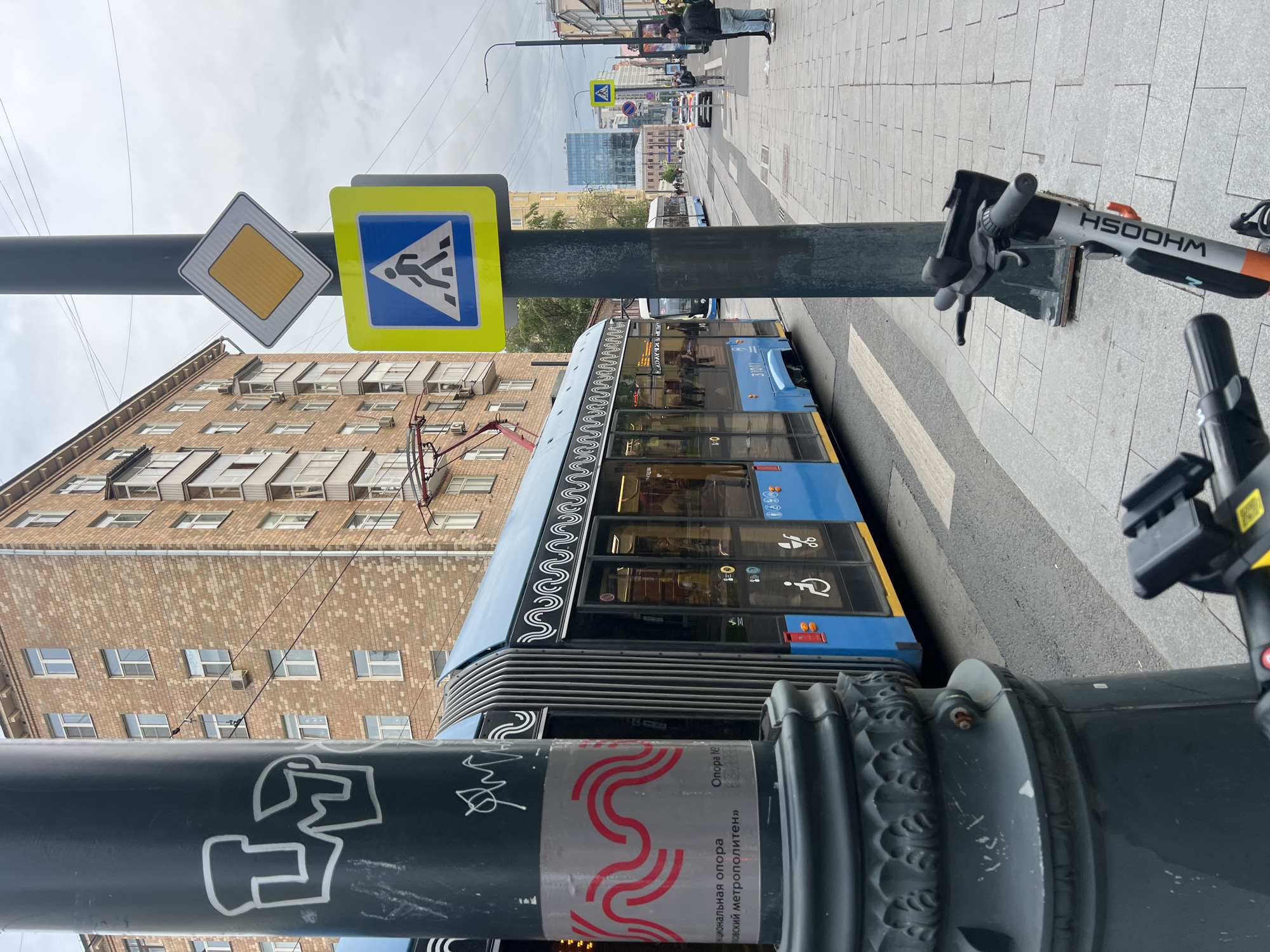 The image, taken from a low angle and rotated counter-clockwise, presents a street scene with several key elements. A blue tram car is positioned on a pedestrian crossing. Street signs including pedestrian crossing and road signs are attached to a lamp post in the foreground. The lamppost is adorned with graffiti and signage. Parked electric scooters are located adjacent to the lamppost on the sidewalk. In the background, there is a building with a brick facade and multiple windows. The street extends into the distance, revealing other buildings and pedestrians. The sky is overcast. The scene is typical of a city street. The location is Unknown, Unknown.