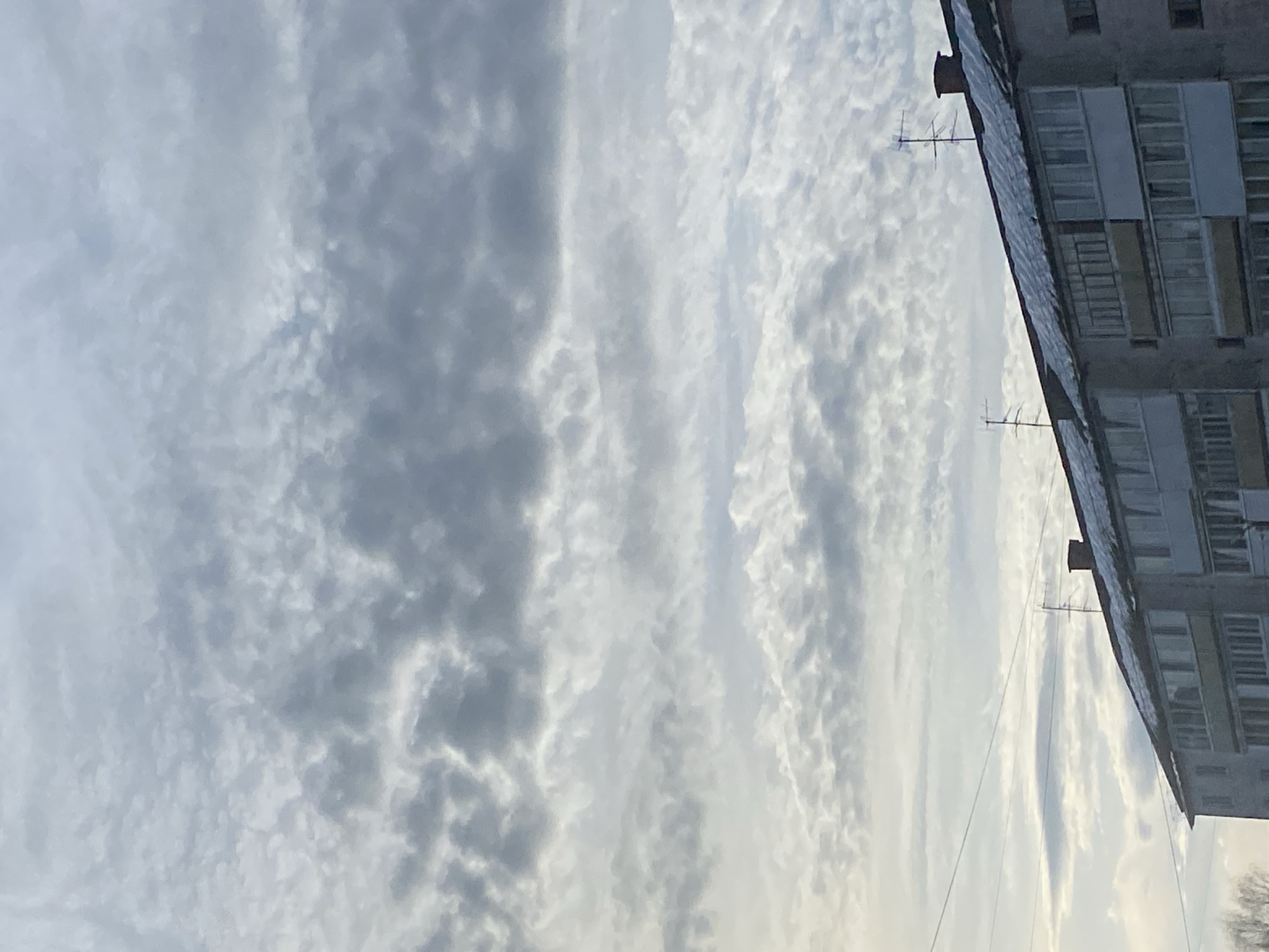 The image depicts an exterior view of a multi-story residential building under a heavily clouded sky in Noviy, Russia. The upper portion of the frame is dominated by a dense, textured cloudscape, primarily consisting of stratocumulus clouds in varying shades of grey and white, with brighter patches indicating diffused sunlight. The lower third of the image features the upper floors and roofline of a concrete-structured apartment building. The building's gabled roof is partially covered with a layer of snow or ice, indicating cold weather conditions. Several television antennas and two brick chimneys are visible along the rooftop. The building's facade is grey, featuring multiple windows and enclosed balconies (loggias). Utility lines traverse horizontally across the mid-ground, above the building. No individuals are visible within the frame. The overall lighting is soft and diffused, characteristic of an overcast day. A few bare tree branches are faintly visible in the extreme lower-left corner.