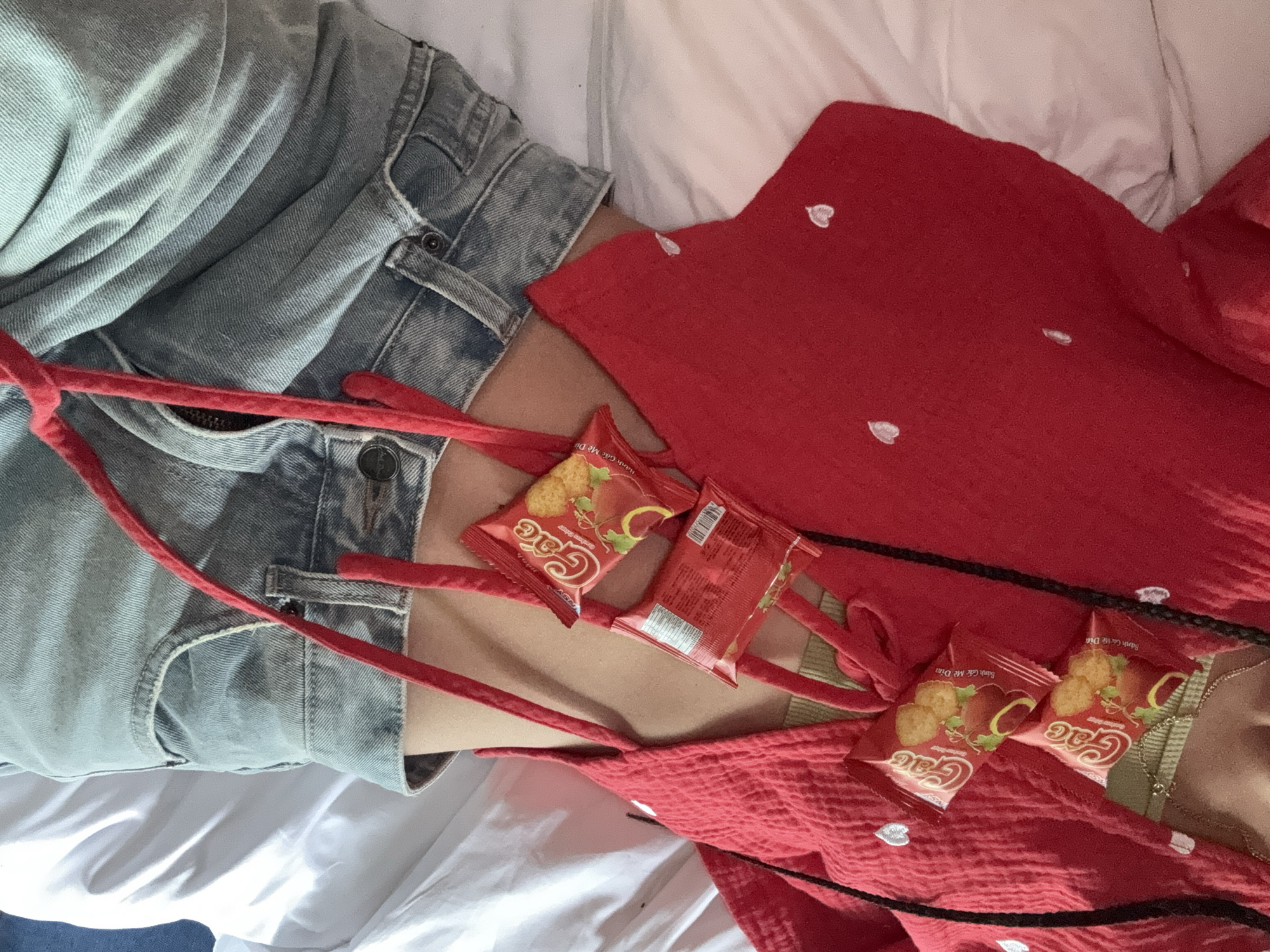 In the city of Mù Cang Chải, Vietnam, an overhead perspective captures a person lying on a white bed sheet, from the mid-torso to the upper thighs. The individual wears light-wash denim shorts and a red, textured garment adorned with small white heart embroidery. Beneath this, a light olive-green bralette and a delicate gold-toned necklace are visible. Red braided cords, either integrated into the red garment or acting as an accessory, are positioned across the upper chest. Five red snack packages, branded "Gác" with images of golden crackers and a reddish fruit, are present; four are face-up on the red garment, held by the cords, and one is reversed, showing a barcode and text. Bare skin is visible above the denim shorts' waistband. The surrounding environment is characterized by rumpled white bedding.