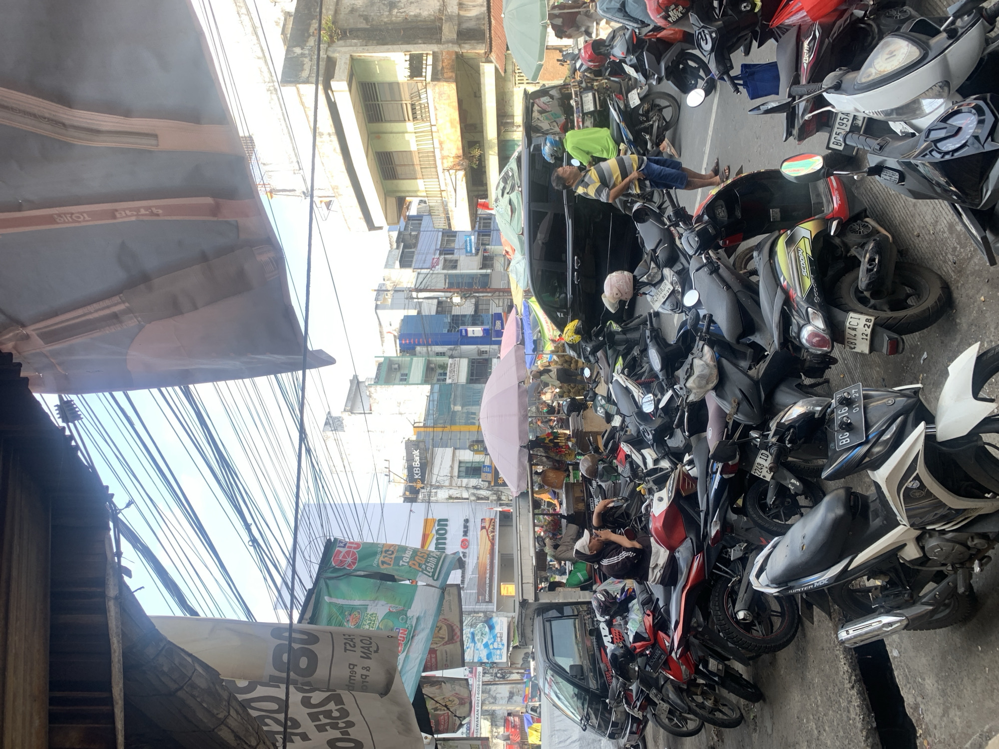 This image depicts a bustling street scene in Palembang, Indonesia, likely a market area based on the variety of goods and the density of people and vehicles. In the foreground, a collection of parked motorcycles and scooters creates a sense of congestion. Several individuals are visible, some seated on motorcycles, one man in a striped shirt speaking on his phone, and others engaged in activities that suggest daily commerce. Behind the motorcycles, a black minivan is parked, partially obscured by the vehicles in front. A pink umbrella is set up, possibly for a vendor, and further back, more stalls and buildings line the street. The architecture appears to be a mix of older and newer structures, with a prominent "KB Bank" sign visible in the background. Overhead, a complex network of wires crisscrosses the sky, indicating a densely populated urban environment. The presence of diverse signage and the style of the vehicles suggest a Southeast Asian setting, with Palembang being a plausible location given its known busy markets and transportation infrastructure.