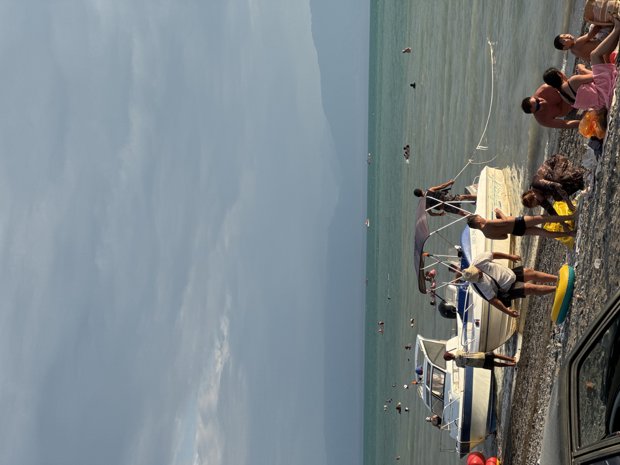 This image depicts a beach scene on a sunny day, likely in the city of Zharbulak, Kazakhstan, with a body of water and a cloudy sky. In the foreground, several people are present on the pebbled shore. To the left, a car is partially visible. Two boats are moored near the shore. One boat, white with blue accents, has a person standing on it, and another person is near it on the shore. The second boat, also white with blue accents, is partially covered by a canopy and has people on it and near it. A yellow and blue inflatable tube lies on the ground. In the middle ground and background, the water is populated with numerous people swimming and enjoying themselves. Several boats are visible further out on the water. The background consists of layered mountains under a sky filled with dramatic clouds.