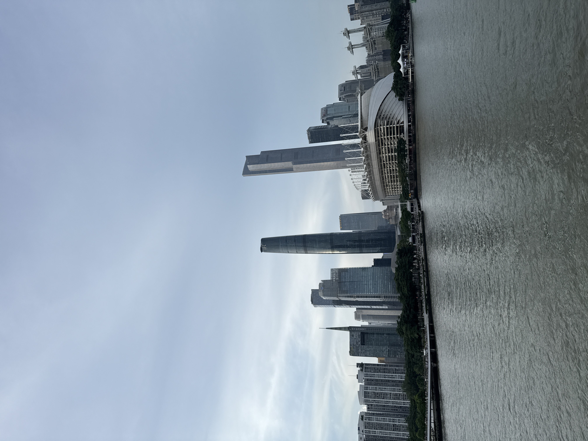 This is a wide-angle, eye-level photograph of the Guangzhou skyline as viewed from across a body of water. The water in the foreground appears to be a river, with slight ripples on its surface and a dark, grayish-green color. A pedestrian walkway lined with trees is visible along the waterfront in the middle ground. Beyond the walkway, a dense collection of modern skyscrapers dominates the background.
Among the prominent buildings are the IFC (International Finance Centre) tower, a tall, slender skyscraper with a dark, reflective facade, and another very tall, angular skyscraper to its left. In the center of the skyline is a distinctive stadium with a white, curved roof structure supported by white beams. To the right of the stadium, there are several other modern buildings, including some with spire-like antenna structures. The sky above is overcast with scattered, wispy clouds, casting a soft, diffused light over the scene. The overall impression is a bustling urban landscape with a focus on modern architecture. This scene is consistent with the view of the Canton Tower and its surrounding skyscrapers in Yuexiu District, China.