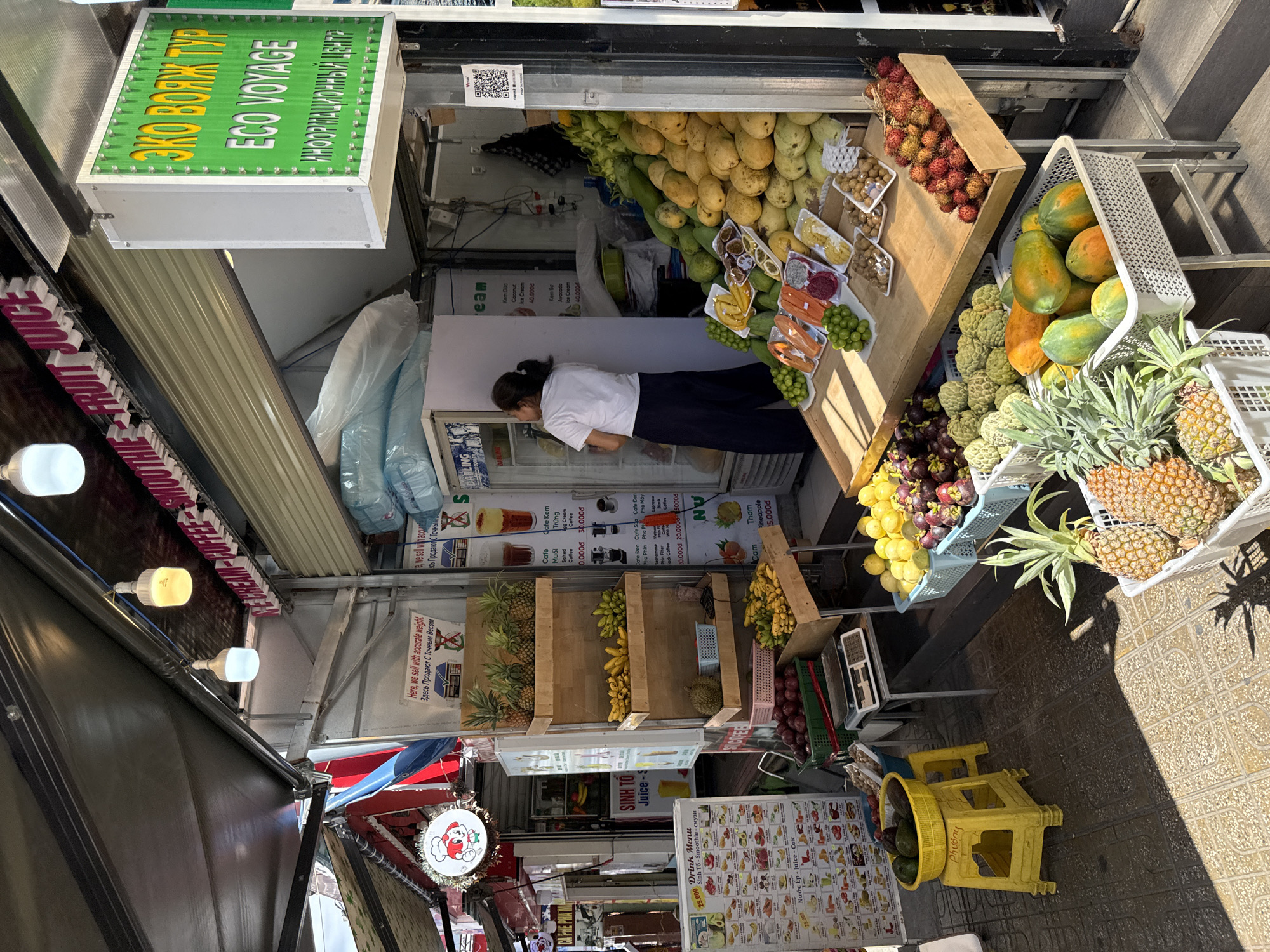 The image shows a fruit stall located on a street in Nha Trang, Vietnam. The stall is an open-air establishment with a canopy providing shade. A variety of fresh fruits are displayed for sale, including pineapples, bananas, mangoes, papayas, rambutan, mangosteen, and other tropical fruits. Some fruits are arranged on wooden shelves, while others are in baskets and crates. A woman wearing a white t-shirt and dark pants is standing inside the stall, facing away from the camera, attending to the produce. Next to the fruit stall, there is a freezer unit with drinks inside, and a sign in Russian that reads "ECO VOYAGE" and "INFORMATION CENTER." A menu board in Vietnamese and English is also visible, listing prices for drinks and fruit items. The street pavement is tiled, and the scene is illuminated by natural daylight and artificial lights hanging from the canopy. The overall impression is that of a bustling street market scene in Vietnam.