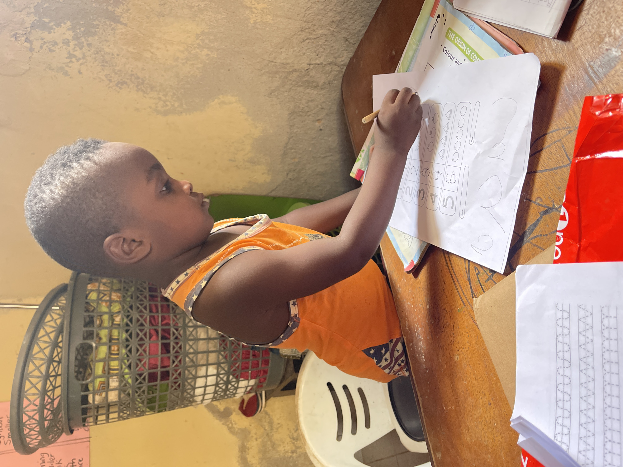 A young child with close-cropped dark hair is sitting at a wooden table in Rido, Nigeria, completing a worksheet. The child, who appears to be a toddler, is wearing an orange top with a stars and stripes design along the edges. He is holding a writing utensil in his right hand, poised to work on a paper covered in geometric shapes and numbers.
The table is cluttered with various papers, including a worksheet titled "THE ORIGIN OF COM". A red package, a box, and a stack of papers are also on the table. The child is sitting on a white plastic chair with cut-out designs. A gray laundry basket filled with clothing is visible behind the child. The wall behind the scene appears to be textured and painted in a muted color.