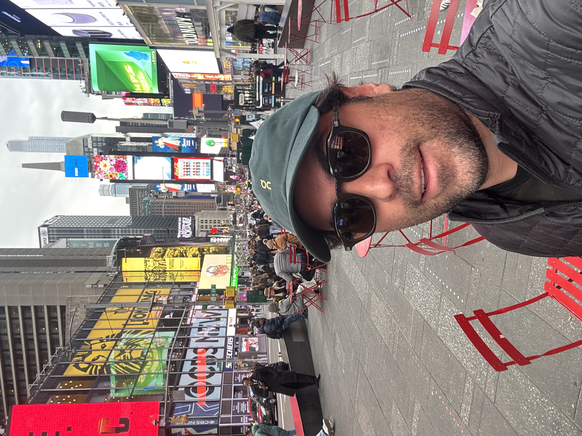 The image depicts a crowded urban scene in Times Square, New York City, United States of America, specifically near West 44th Street.
In the immediate foreground, a male subject is taking a selfie. He wears a dark green baseball cap with a gold "oc" logo, black sunglasses reflecting a partial American flag, a black quilted jacket, and has a short beard. His mouth is slightly open in a subtle smile. Several red metal outdoor chairs are visible around him.
The mid-ground features a paved pedestrian area with multiple red metal tables and chairs, some occupied by small groups of people sitting and appearing to converse. Numerous pedestrians are walking throughout the square. One individual in a dark trench coat stands near a planter box, holding a mobile device.
The background is dominated by towering buildings adorned with numerous large, illuminated digital billboards displaying advertisements, including "CBS NEWS," "SKECHERS," "TISSOT," "OLD NAVY," and "THE LION KING MUSICAL." Traffic is visible on the street beyond the pedestrian area. The sky is overcast, suggesting daytime with subdued lighting.