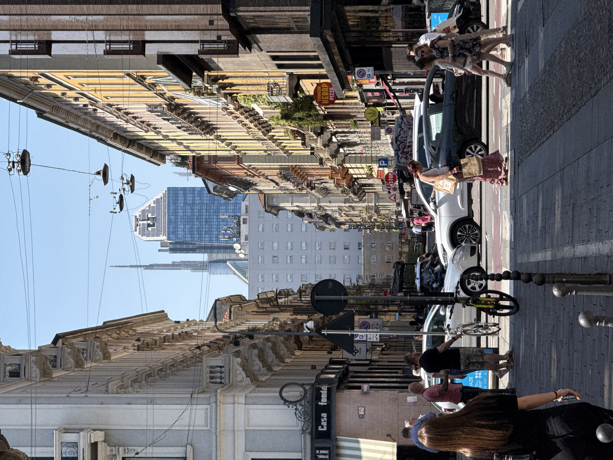 The image shows a street in Milan, Italy. The street is lined with buildings of varying heights, constructed in architectural styles including classical and modern. Pedestrians occupy the sidewalks on both sides of the street, including groups of people walking and individuals interacting with parked vehicles. Several cars are parked alongside the street. A bicycle is leaning against one of the vehicles. Street signage is visible on the sides of buildings. The sky is clear and the lighting appears to be mid-day sun. In the background, there are modern skyscrapers.