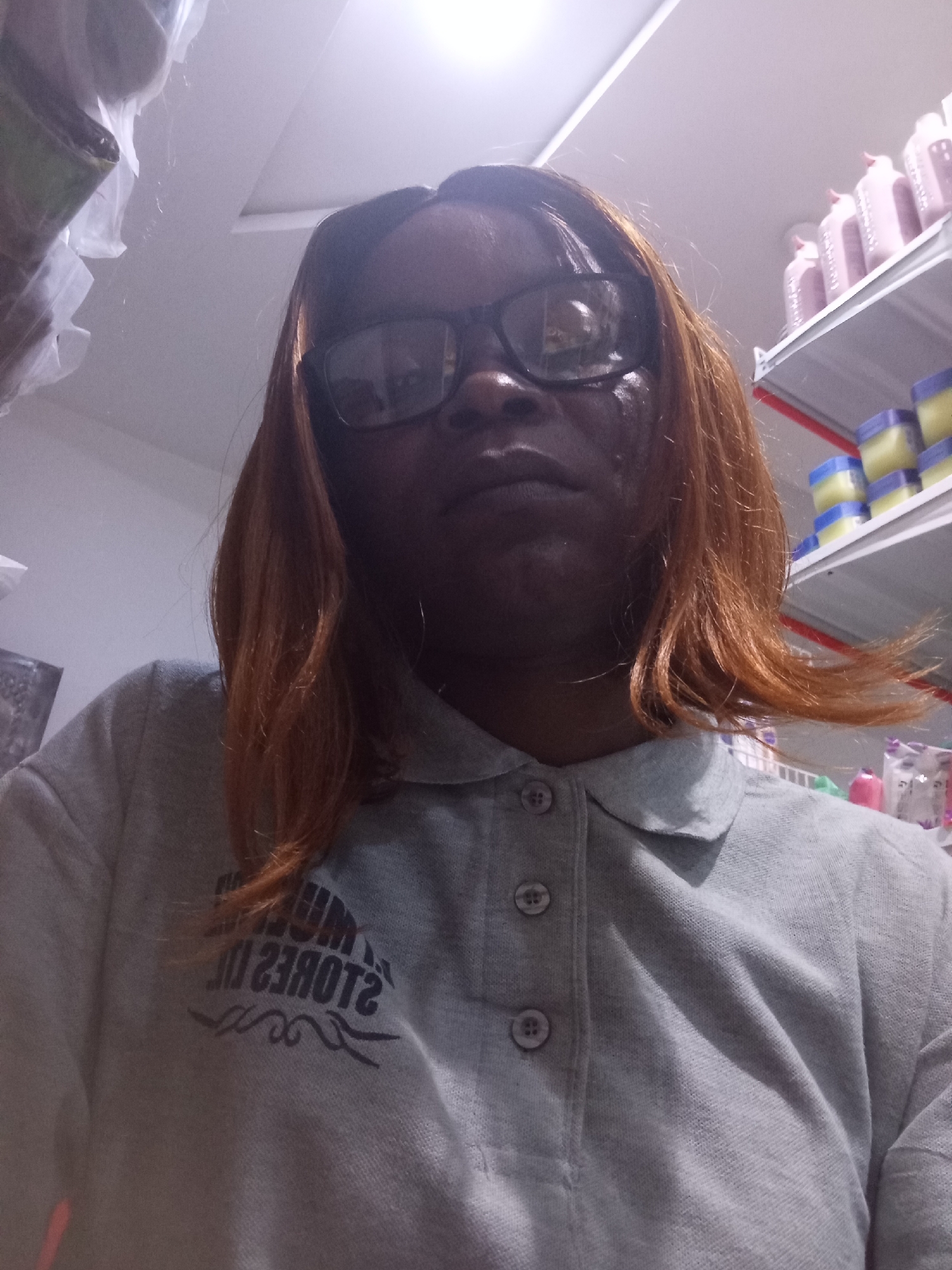 A woman with auburn hair and glasses is taking a selfie. She is wearing a grey polo shirt with a black logo on the left side. The logo appears to be text that is partially obscured and reversed, with a decorative element below it. She is standing in front of shelves stocked with various products, including bottles and jars. The lighting suggests an indoor environment, possibly a retail store. There is no specific information in the image to indicate the location in the city of Howayi, Nigeria.