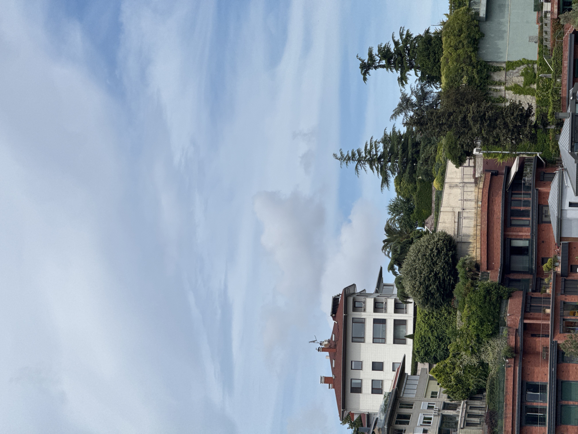 This is a vertical, eye-level, medium shot of a residential area in Beşiktaş, Turkey. The sky is a soft blue with scattered white clouds, suggesting it is daytime. Lush green trees and vegetation are interspersed with modern multi-story buildings constructed from brick and white stucco. Some buildings have large windows and balconies, hinting at a contemporary urban setting. The image captures a serene moment, with no visible people or specific activities taking place. The overall impression is one of a well-maintained, affluent neighborhood with a strong integration of nature and architecture.
