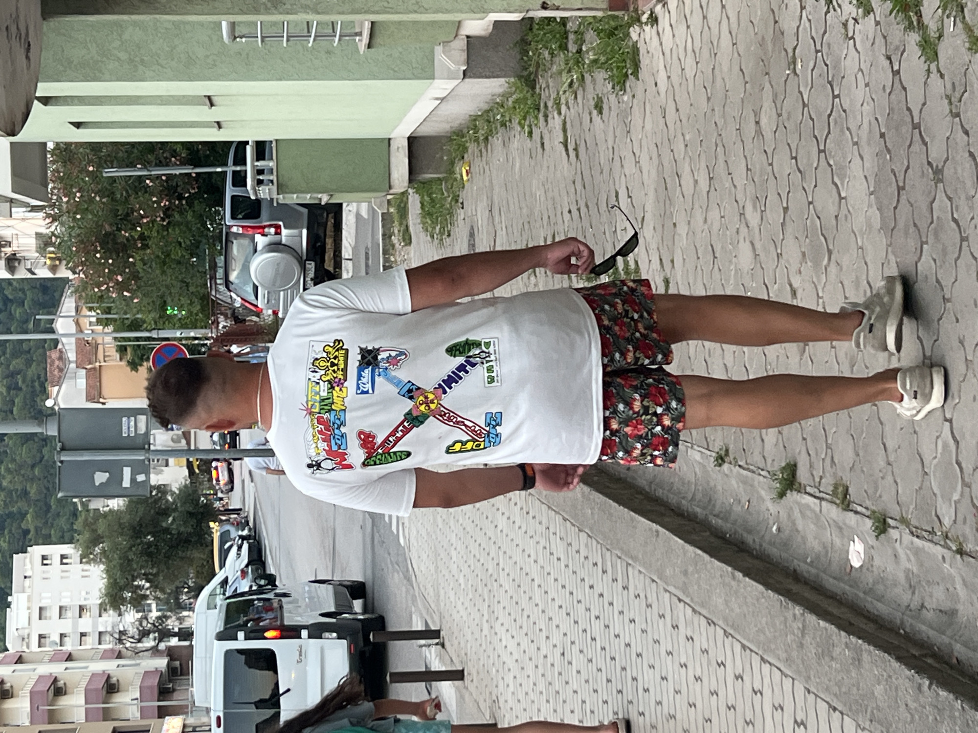 In Budva, Montenegro, a man with a fade haircut walks away from the camera on a paved sidewalk. He is wearing a white t-shirt with colorful graphics and floral patterned shorts. He holds a pair of sunglasses in his right hand. To his left, a drain runs along the edge of the sidewalk, separating it from a grassy area. Across the street, vehicles are parked and moving, including a white van and a silver SUV. Buildings line the street, with apartment buildings visible in the background. The overall scene depicts a street in a European coastal town.