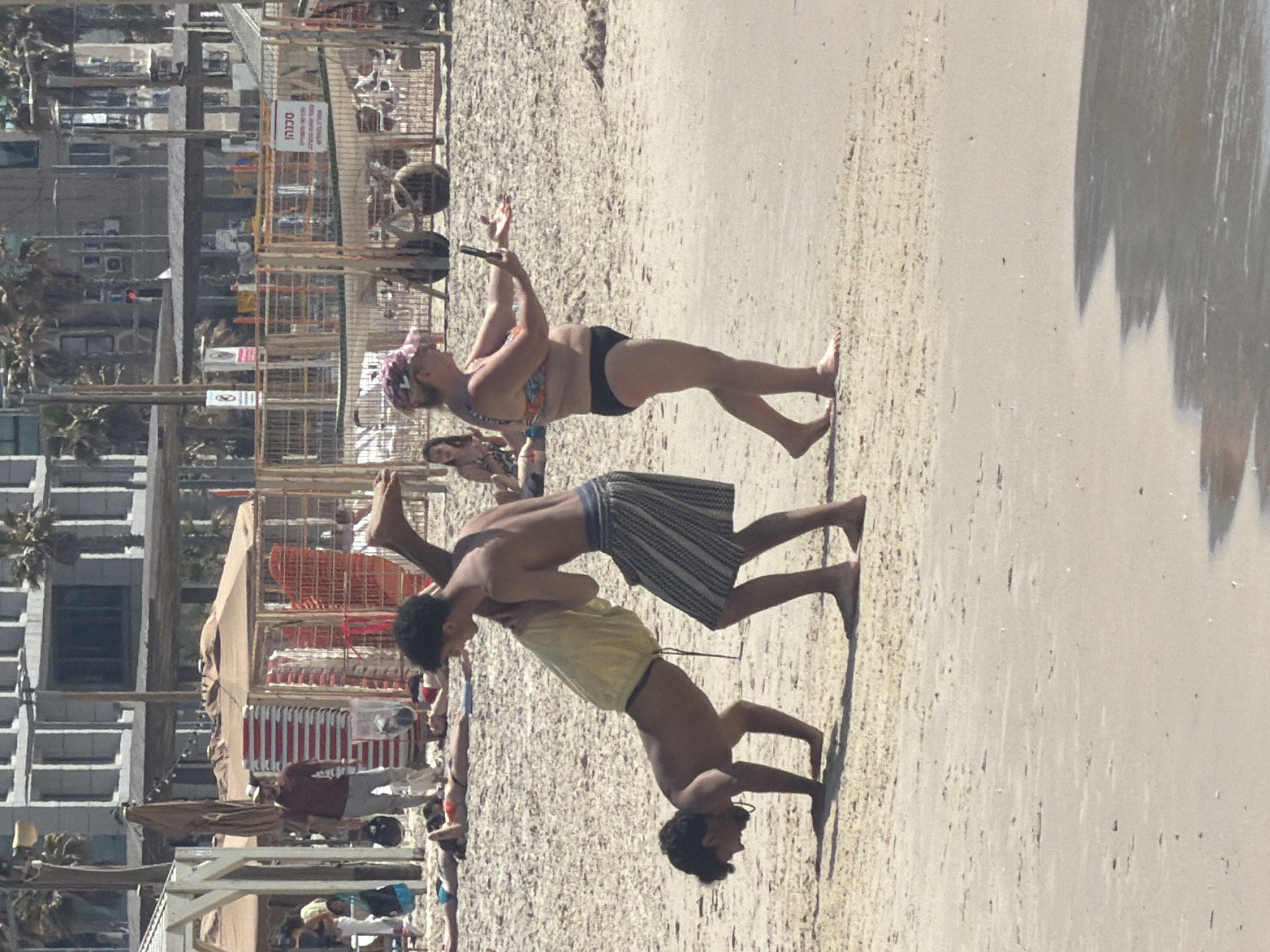 The image shows a crowded beach in Tel-Aviv, Israel. The beach is sandy with a mix of coarse and fine grains. In the foreground, two young men are performing a balancing acrobatic move, with one supporting the other in a headstand position on the sand. Behind them, a woman in swimwear holds a phone, seemingly taking a picture of the acrobatic act. Other people are scattered on the beach, some sunbathing and others walking around. Buildings are visible in the background, indicating an urban setting adjacent to the beach. Beach infrastructure, such as umbrellas and chairs, is also present. The lighting suggests it is daytime. The city of Tel-Aviv is not identified in this image.