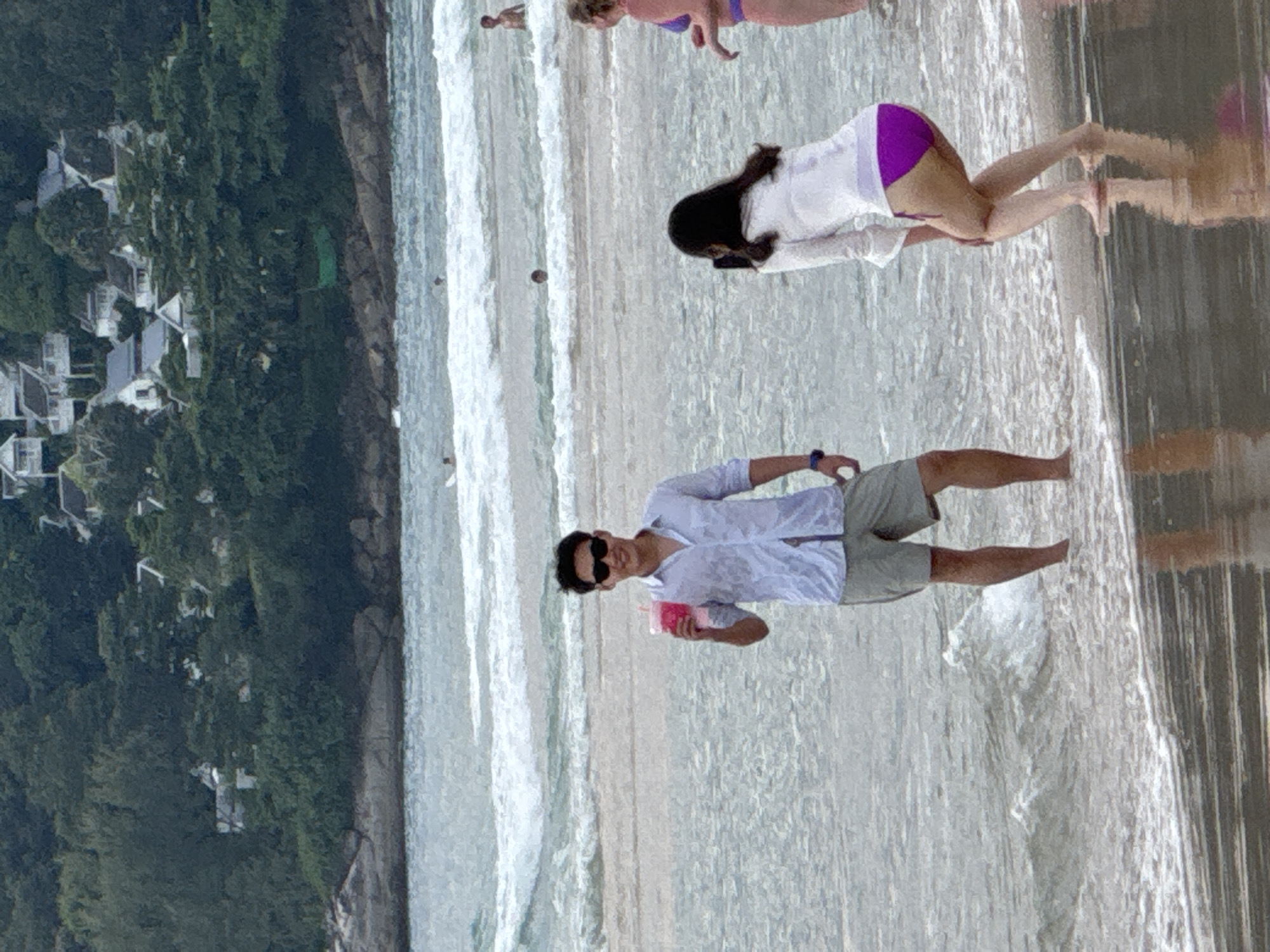 In Karon, Thailand, a man in sunglasses and a light blue open-collared shirt stands in shallow water holding a pink drink. He wears grey shorts. To his right, a woman in a white shirt and purple bikini bottoms squats down, looking at her phone. Further to the right, a woman in a purple bikini is partially visible. The background shows the ocean with waves and a lush green hillside dotted with white buildings. The wet sand reflects the sky and surrounding elements.
