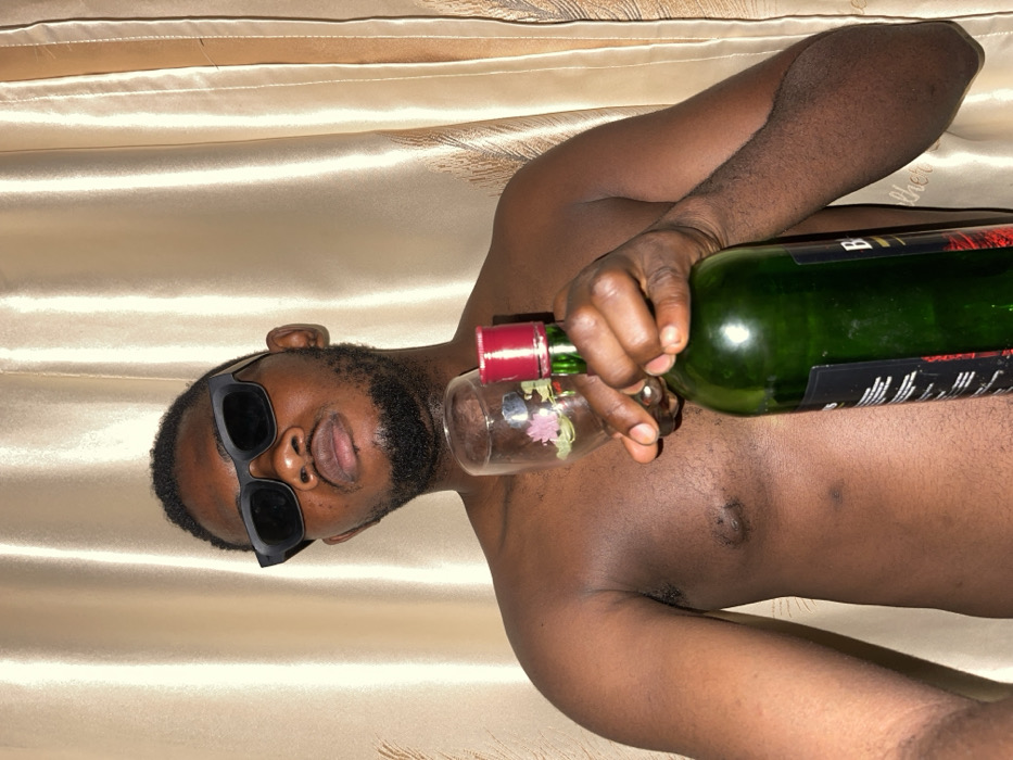 A shirtless man with dark skin, wearing sunglasses, is lying on his side on a shiny, gold-colored surface. He is holding a green wine bottle with a red top and pouring the wine into a clear glass with floral designs. The bottle label is partially visible, featuring white lettering. The man has a beard and appears relaxed. His torso and upper arm are visible against the background.