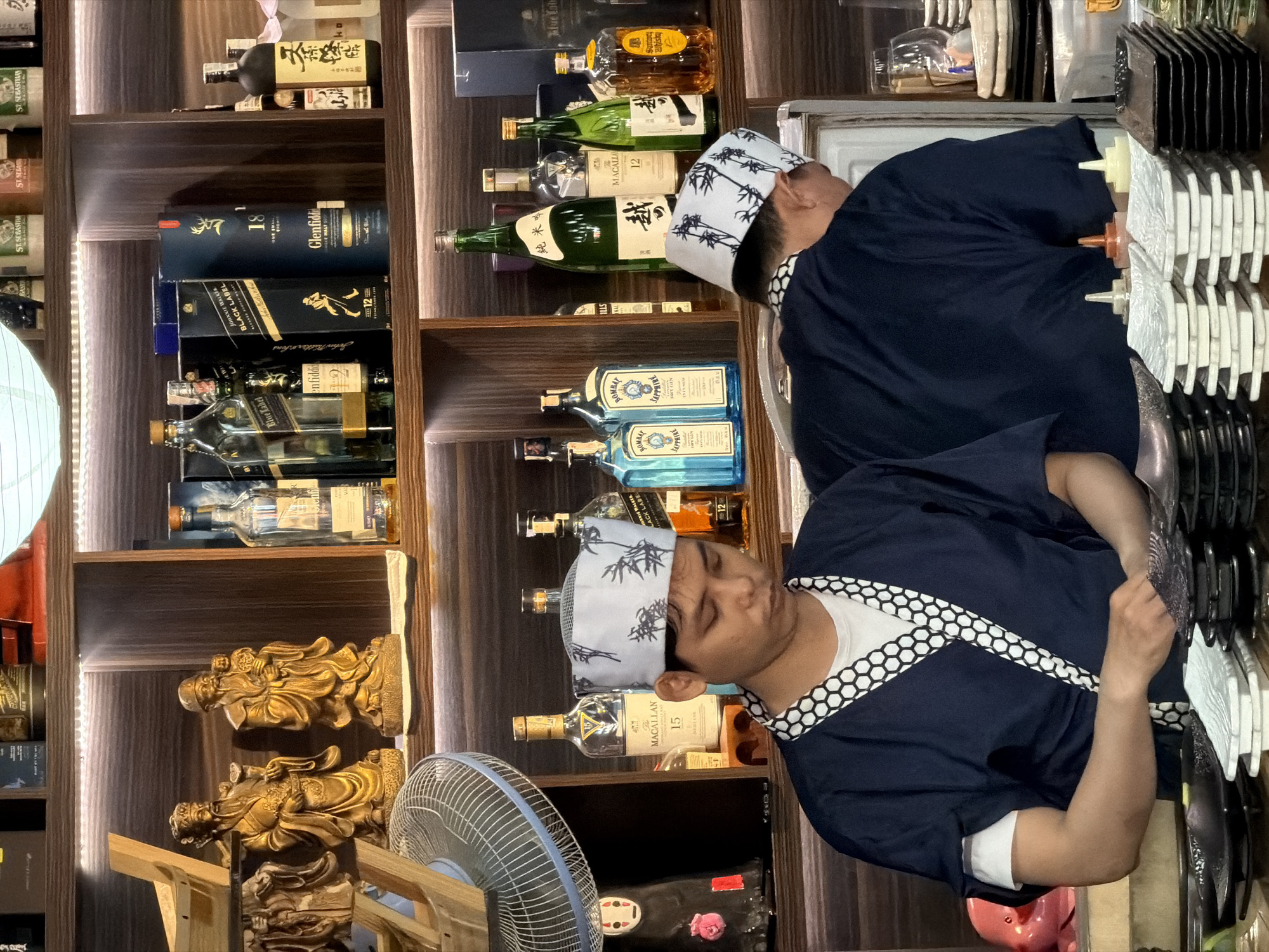 The image depicts the interior of a bar or restaurant in Nha Trang Ward, Vietnam. The scene is set against a backdrop of wooden shelves illuminated by warm ambient lighting, displaying an assortment of alcoholic beverages, primarily whiskey and gin bottles. On the shelves, there are also decorative items including golden statues of men and a few packaged boxes.
In the foreground, two individuals, presumably staff members, are visible. Both are wearing traditional dark blue Japanese-style work attire with white patterned trim around the collars and hats. One individual, facing the camera and slightly to the left, is looking down, seemingly engaged in preparing something. The other individual, to the right, is seen from the back, also appearing to be working behind a counter.
The counter area in front of them is cluttered with stacks of plates, small condiment bottles, and other serving ware, suggesting an active food or drink service environment. To the left, a blue-and-white fan is partially visible. The overall impression is one of a well-stocked establishment with a focus on spirits, possibly a Japanese restaurant or a bar with a significant Japanese whisky selection.