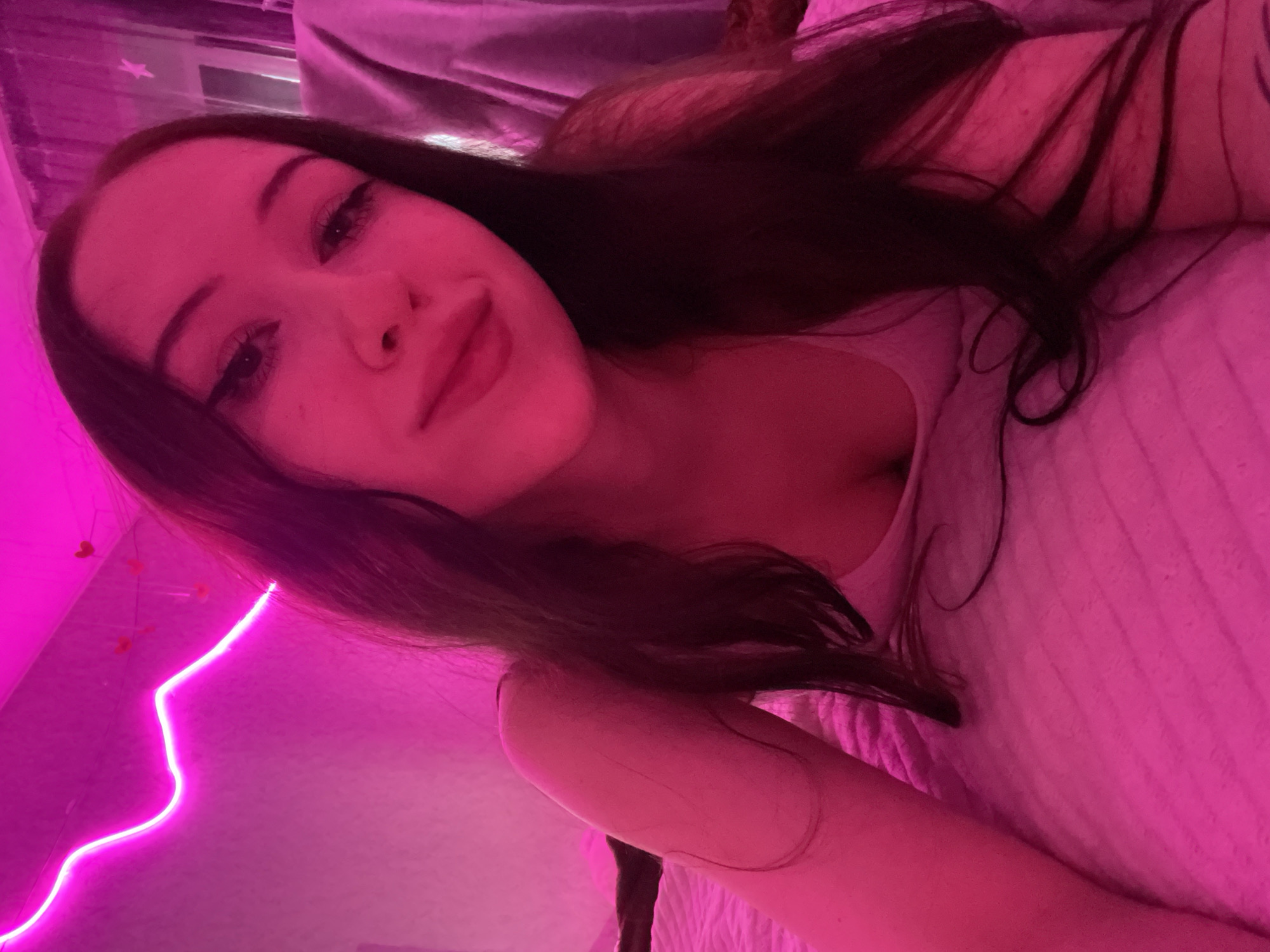 In a dimly lit room, a young woman with long, dark hair and prominent eye makeup lies on a pink bedspread. A neon pink light fixture snakes along the wall behind her, casting a vibrant glow over the scene. Her face is tilted towards the camera, with a slight smile playing on her lips. The room appears to be a bedroom, with a curtained window visible in the background. The overall impression is one of intimacy and a youthful, playful atmosphere. The image has not been geolocated to Votkinsk, Russia.