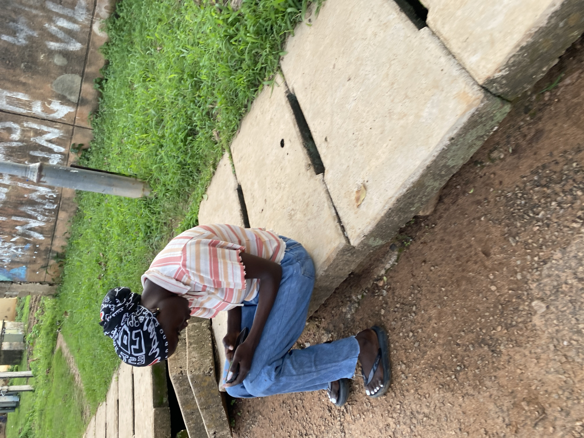 A young Black female is seated on concrete slabs, looking down at a smartphone in her hands. She is wearing a black and white patterned headwrap, a horizontally striped t-shirt with peach, white, and red stripes, and light blue jeans. Her feet are clad in black flip-flops, and her toenails are painted white. The concrete slabs are laid out in a path, with some appearing broken or uneven. To the left of the slabs is a dirt path. To the right and behind the slabs is a grassy area, and beyond that, a textured wall with graffiti. A grey metal pole stands near the wall. The scene appears to be outdoors in a potentially urban or semi-urban environment, possibly in Rafin Par, Nigeria, given the context.