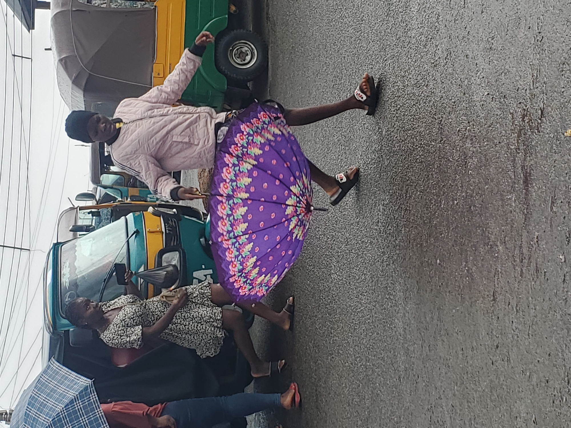 A person in a pink jacket and black beanie holds a vibrant purple umbrella with a floral pattern. They are standing with one leg extended, mid-stride, on a wet, grey asphalt road. The person wears black slides with white soles. Behind them, a green and yellow auto-rickshaw, a common mode of transport, is visible. Further back, another auto-rickshaw is parked. Two women are seated inside one of the auto-rickshaws. One woman, wearing a black and white patterned dress, holds a smartphone. A person in a red top and blue jeans lies down, partially covered by a blue and white checkered umbrella. The scene suggests it is raining, as the ground is wet and umbrellas are in use. The sky appears overcast, indicating a cloudy day, likely daytime due to the presence of auto-rickshaws and people active in the street. The overall atmosphere is a typical street scene in Gwong, Nigeria, during wet weather.