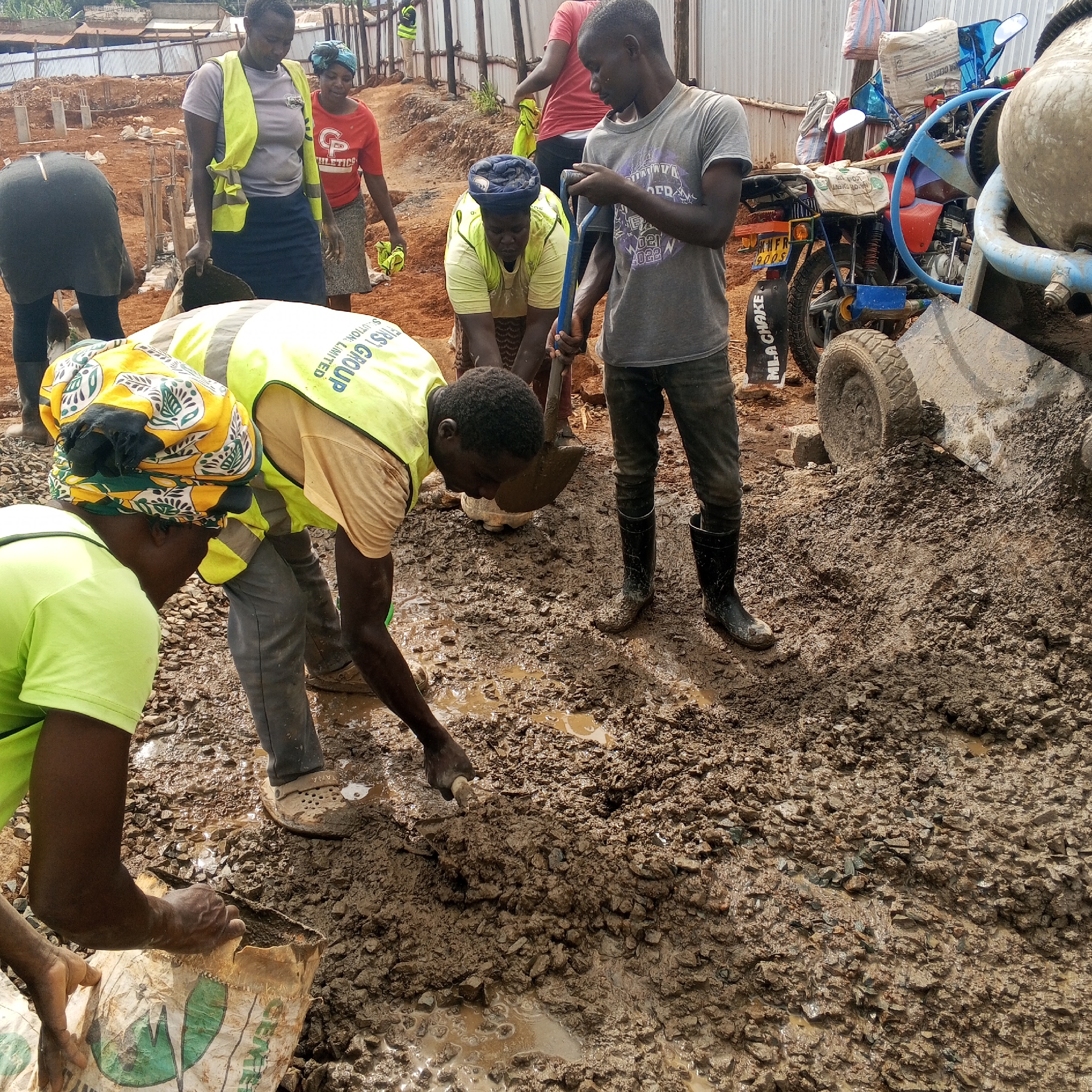 The image depicts a muddy construction site, possibly in Borabu Chitago ward, Kenya, based on visual context and implied details from limited visible text. Several individuals are engaged in labor, including mixing cement. Some wear yellow safety vests, while others are in casual attire. One person wields a shovel, and another appears to be opening a bag labeled "CEMENT." A cement mixer and a motorcycle are partially visible, suggesting their use in the construction process. The background shows unfinished structures and scaffolding. The overall environment is unpaved, with muddy terrain dominating the foreground.