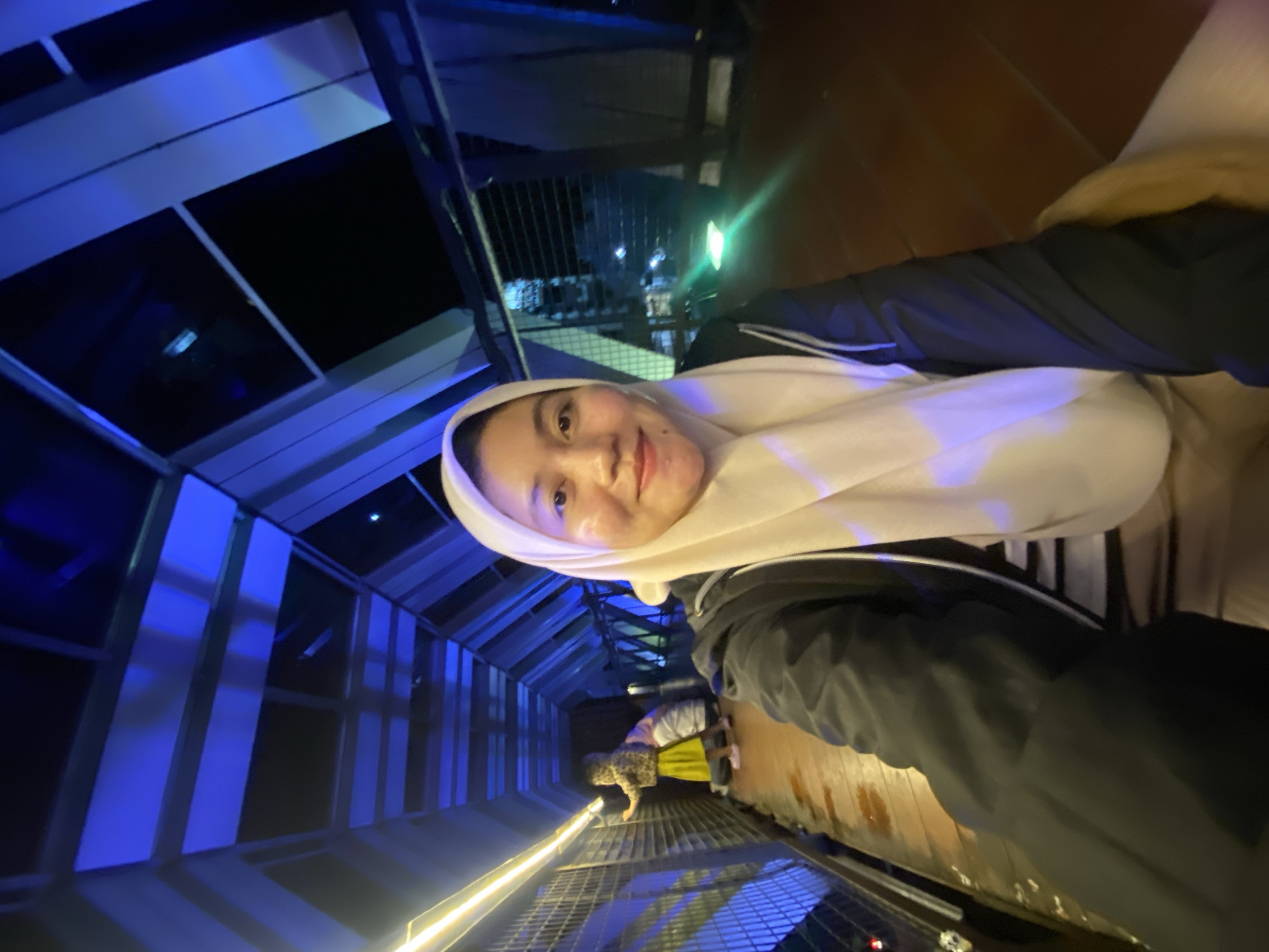 The image shows a woman wearing a light-colored hijab and a black jacket, taking a selfie. She is in a long, narrow corridor with blue lighting on the left and a wooden floor on the right. The corridor appears to be part of a building or a walkway with metal railings on both sides. In the background, another person is visible further down the corridor, wearing a leopard-print jacket and a yellow skirt. The lighting suggests it is nighttime or an indoor environment. The woman's expression is a soft smile. The location is identified as Banjarbaru, Indonesia.