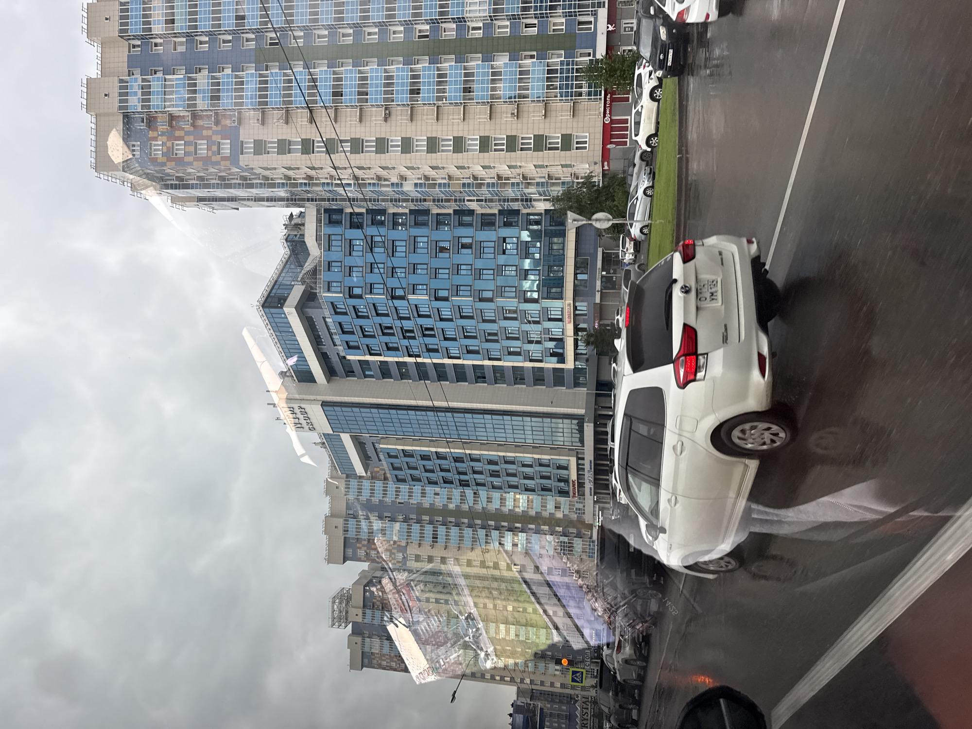 The image depicts a street scene in Krasnoyarsk, Russia, characterized by modern, multi-story buildings and traffic. In the foreground, a white station wagon is driving on a wet road, reflecting the surrounding environment. The buildings are tall, with facades featuring a combination of glass panels, colored bricks, and concrete. The sky is overcast, suggesting cloudy weather. Several other vehicles are visible on the road and parked along the side. The overall impression is that of an urban environment with ongoing traffic flow.