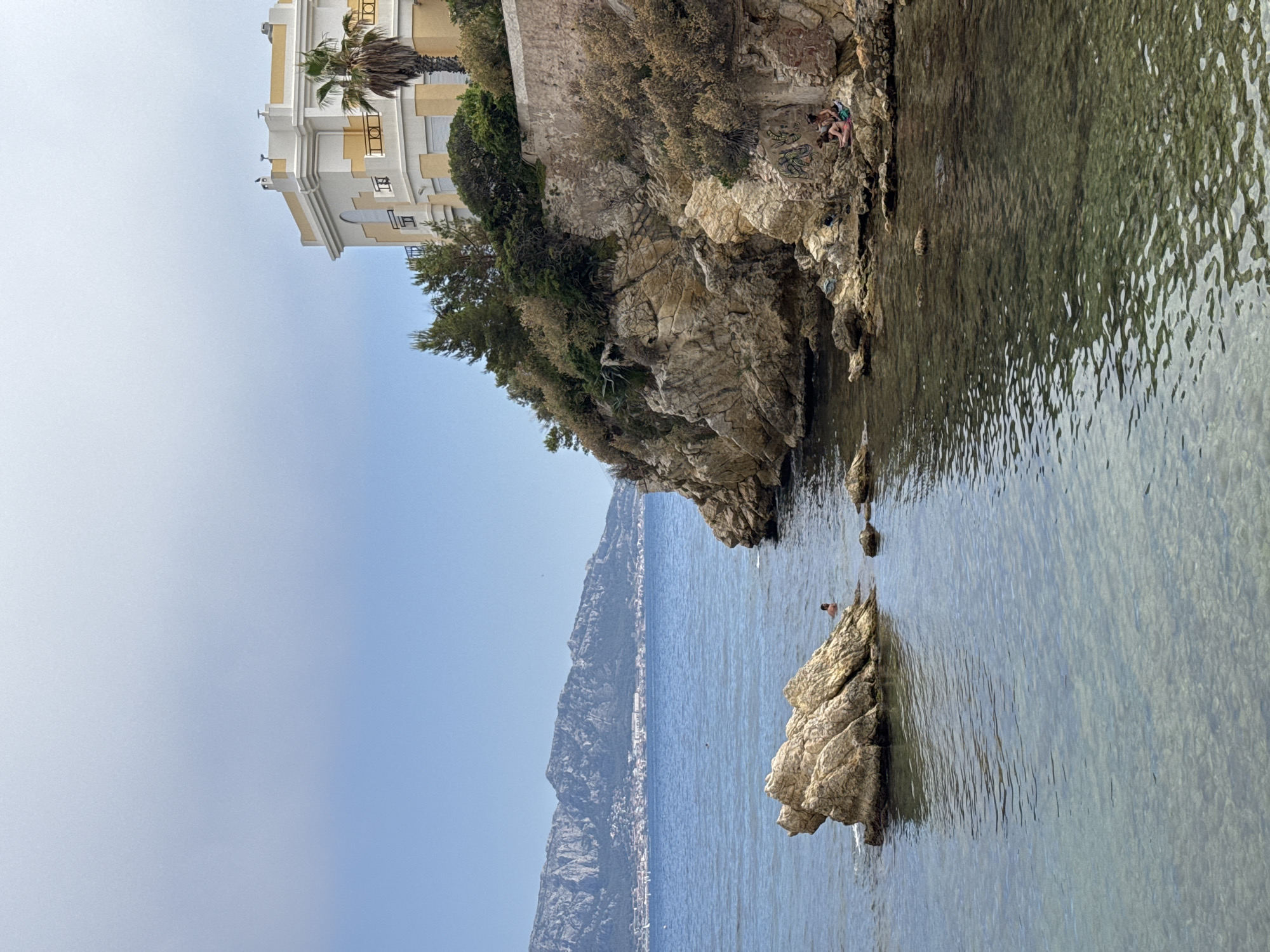 The image shows a view of the Mediterranean Sea in Marseille, France, with a rocky coastline in the foreground and a city skyline in the distance. On the right side of the image, a pale yellow and white building sits atop a cliff, surrounded by lush green trees and shrubs. The building has a balcony with yellow railings and a palm tree is visible to its right. The rocky cliff face is textured and shows signs of erosion and vegetation growth. In the water, there are several large rocks. On one of these rocks, a person is swimming. Further to the right, on the rocky shore, two people are sitting, one wearing a pink bikini and the other wearing a green garment. The water is clear and calm, with sunlight reflecting off its surface. In the background, a chain of mountains stretches across the horizon, with a city built along its base. The sky is a light blue with a few wispy clouds. The overall scene depicts a serene coastal landscape with elements of both nature and human habitation.
