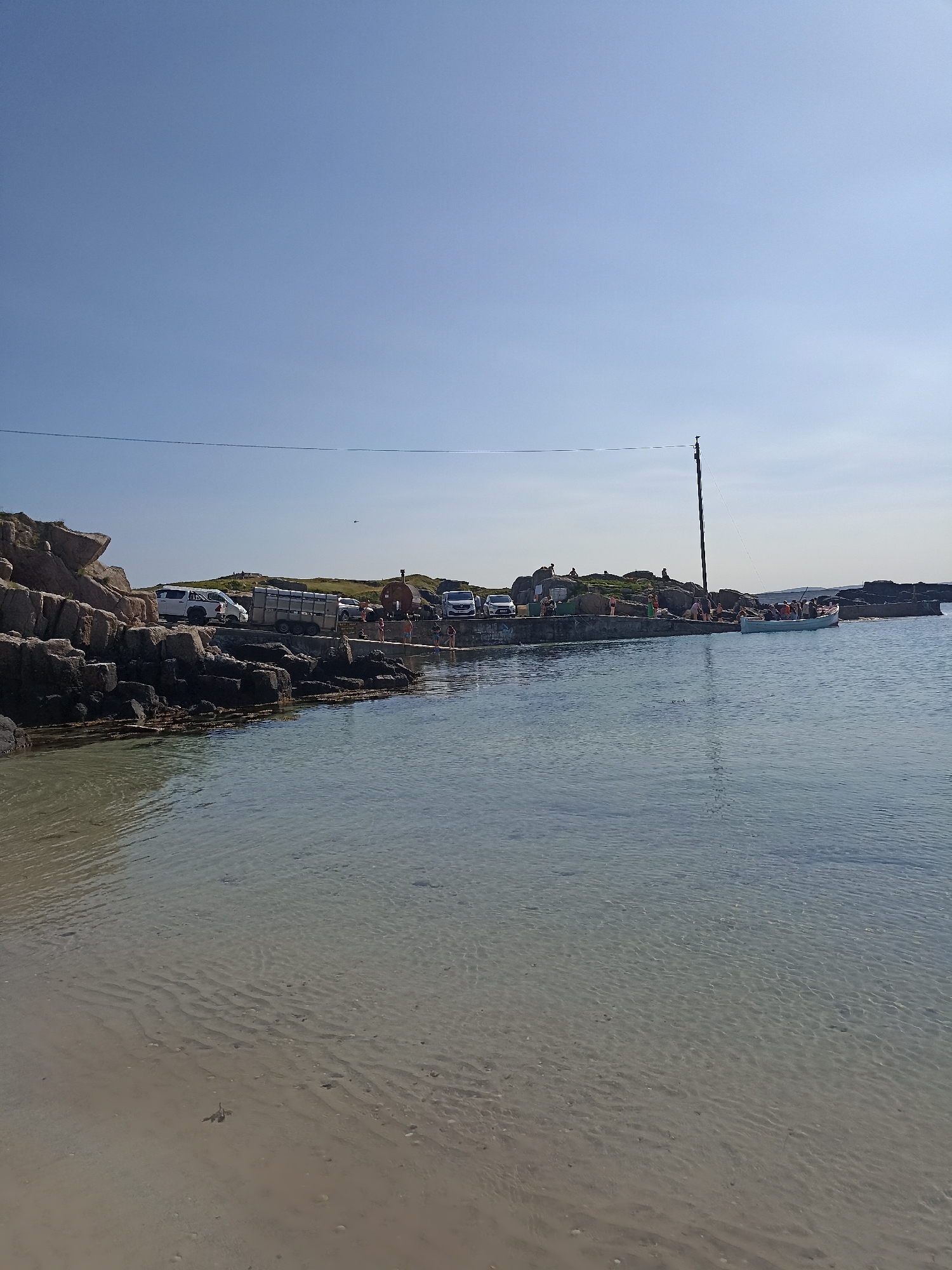 The image captures a bright, sunny day at a coastal location in Carnboy, Ireland. The foreground features a sandy beach with clear, shallow water rippling over the seabed, revealing sand patterns and scattered shells. To the left, a rocky shoreline extends into the frame, with dark, textured rocks and some patches of seaweed.
Beyond the rocky edge, a concrete pier or walkway runs parallel to the water. Several vehicles are parked along this pier, including a white van, a car trailer, and other cars. A few individuals are visible on the pier, some appearing to be standing or walking.
Further along the pier, a small, light-blue boat is docked, with several people on board. A tall wooden pole with wires attached stands near the boat, extending upwards into the clear blue sky. To the right of the boat and further along the pier, there are more rocks and a small, round, brown structure that appears to be some sort of building or shelter. The background shows a hilly landscape with green vegetation under a vast, cloudless sky. The overall scene suggests a casual, possibly recreational, setting by the sea.