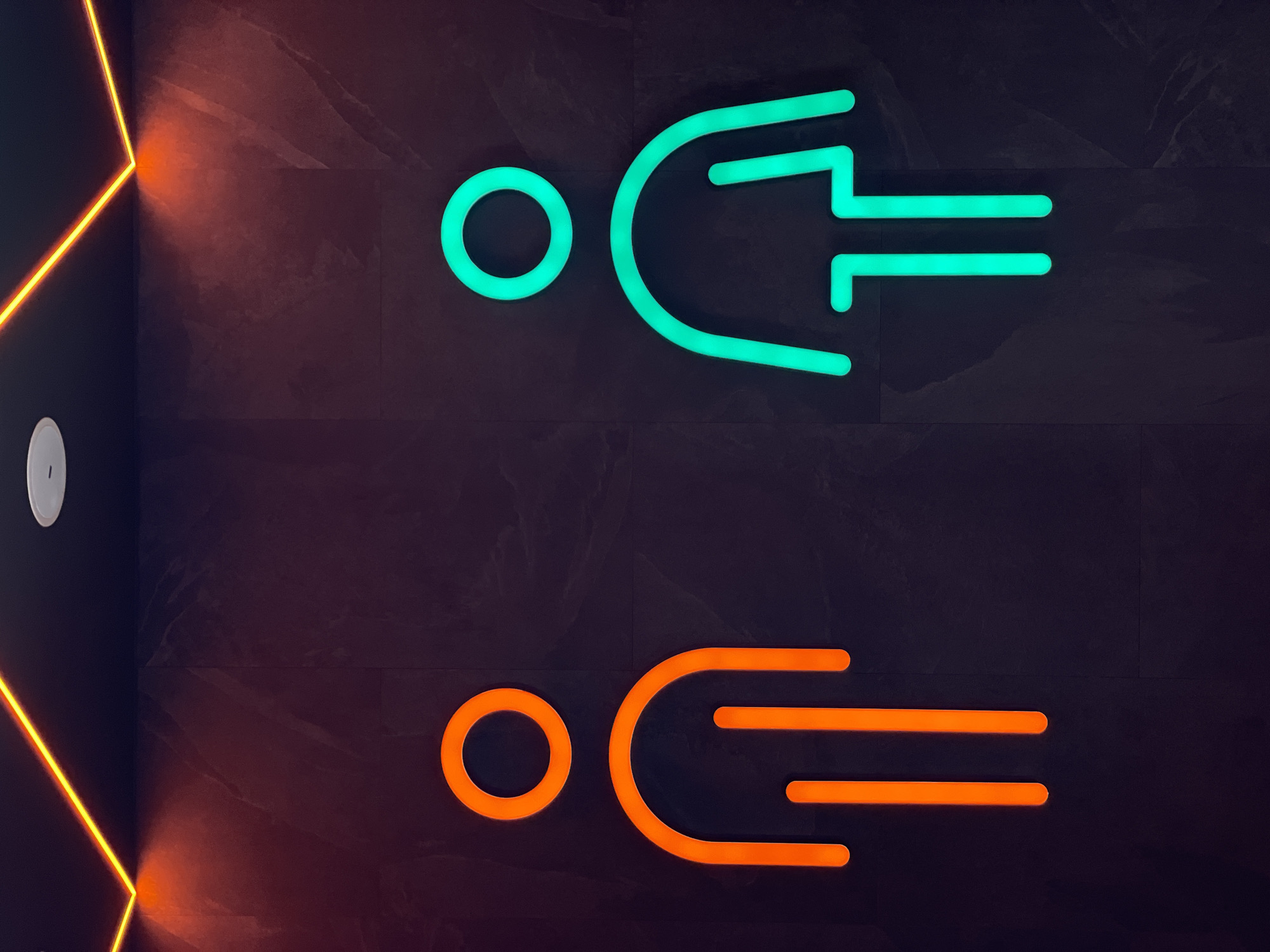 The image shows illuminated restroom signs on a dark, textured wall. The top sign is green and appears to represent a stylized figure with a head (circle) and a body with three horizontal lines extending from a curved shape. The bottom sign is a similar design but in orange. The background wall is dark with subtle patterns, suggesting a tile or stone material. To the left, a wall corner is visible with a vertical strip of orange neon lighting. A small, round, white object (likely a sensor or dispenser) is also on the left wall. This image was taken in Kazan, Russia.