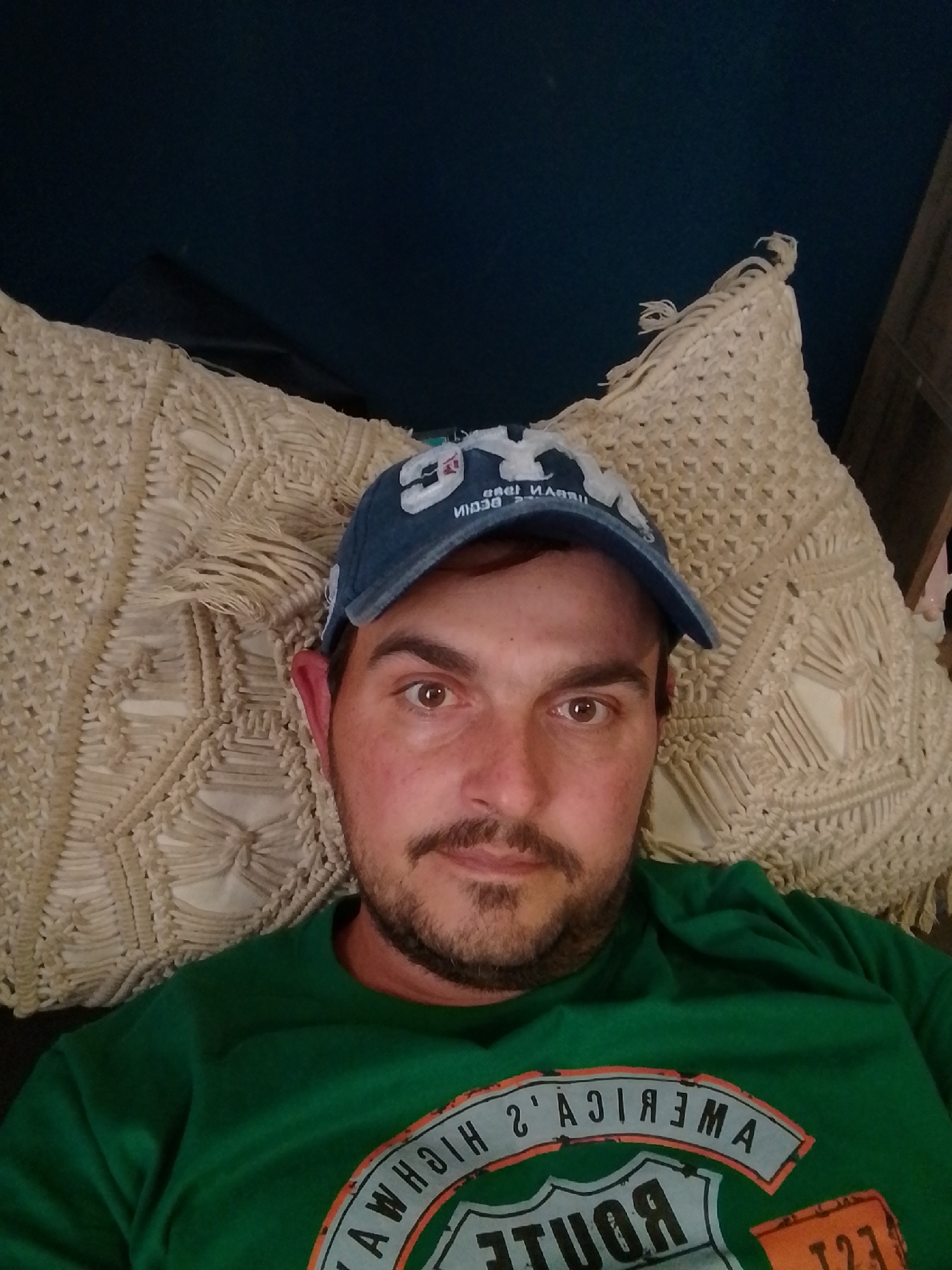 A medium shot, taken from a high angle, shows a man wearing a blue baseball cap and a green t-shirt. He is leaning back against a cream-colored, woven cushion. The man has brown hair and a mustache, and he is looking directly at the camera with a neutral expression. The t-shirt has a graphic on it that appears to be related to a route or highway. The background is dark blue, suggesting an indoor setting. The image does not provide any information about the location in the city of Wedel, Germany.
