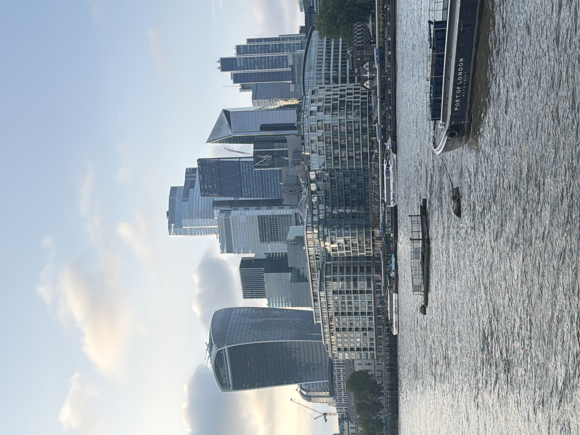 This image depicts a view of the city of London, United Kingdom. Several modern skyscrapers dominate the skyline, including the "Walkie Talkie" building. The River Thames is visible in the foreground, with a large boat, marked "Port of London Authority," on the water. The scene appears to be captured during the daytime, with a partly cloudy sky.
