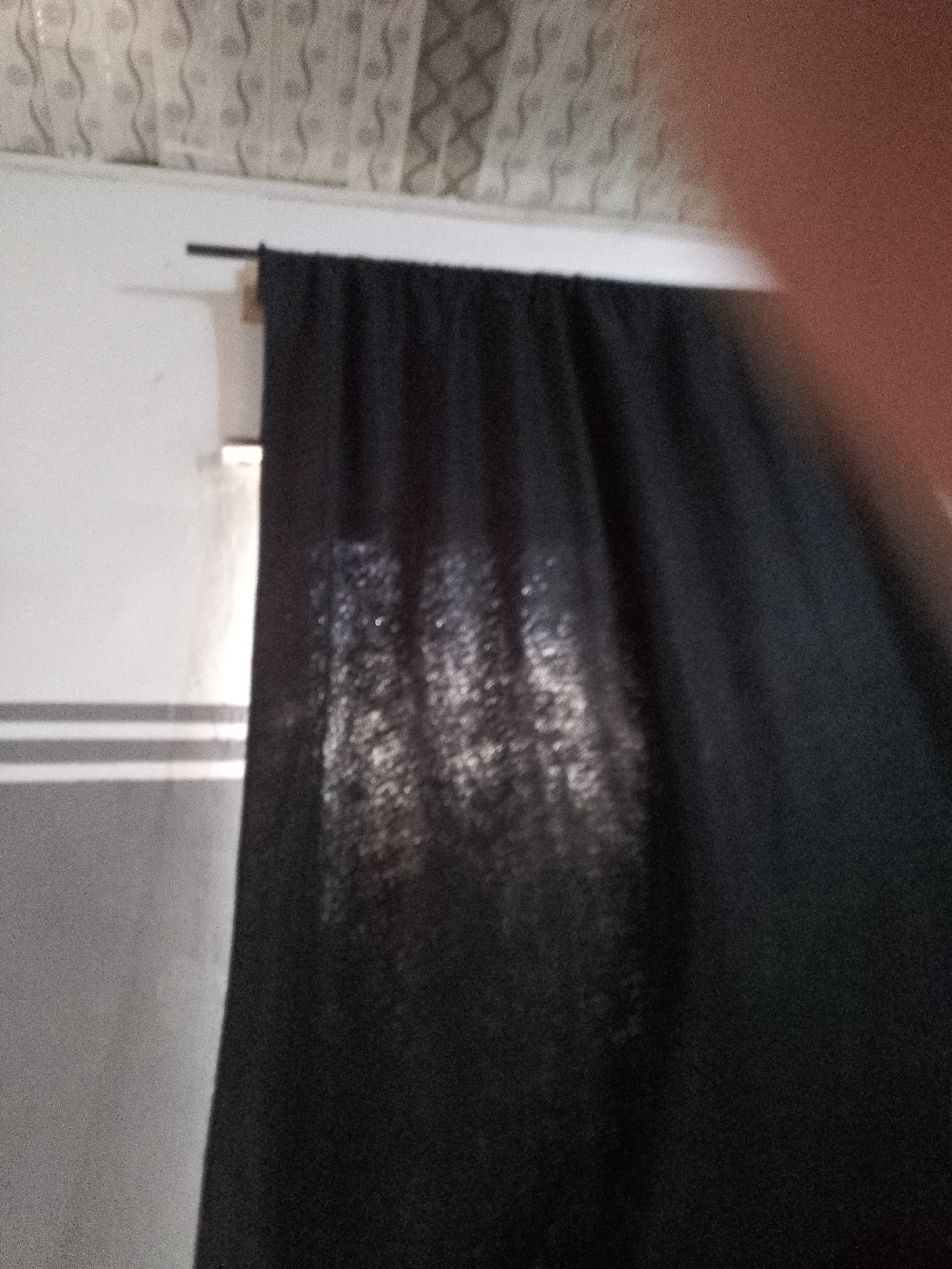 The image depicts a close-up view of a dark curtain hanging from a rod. The curtain has a textured appearance with some light filtering through it, creating a shimmering effect. To the left of the curtain, a white wall is visible, with some horizontal grey lines. Above the wall and curtain, a textured ceiling is partially visible. The scene appears to be indoors, possibly in a residential or commercial setting. No people or specific locations within a city are identifiable.