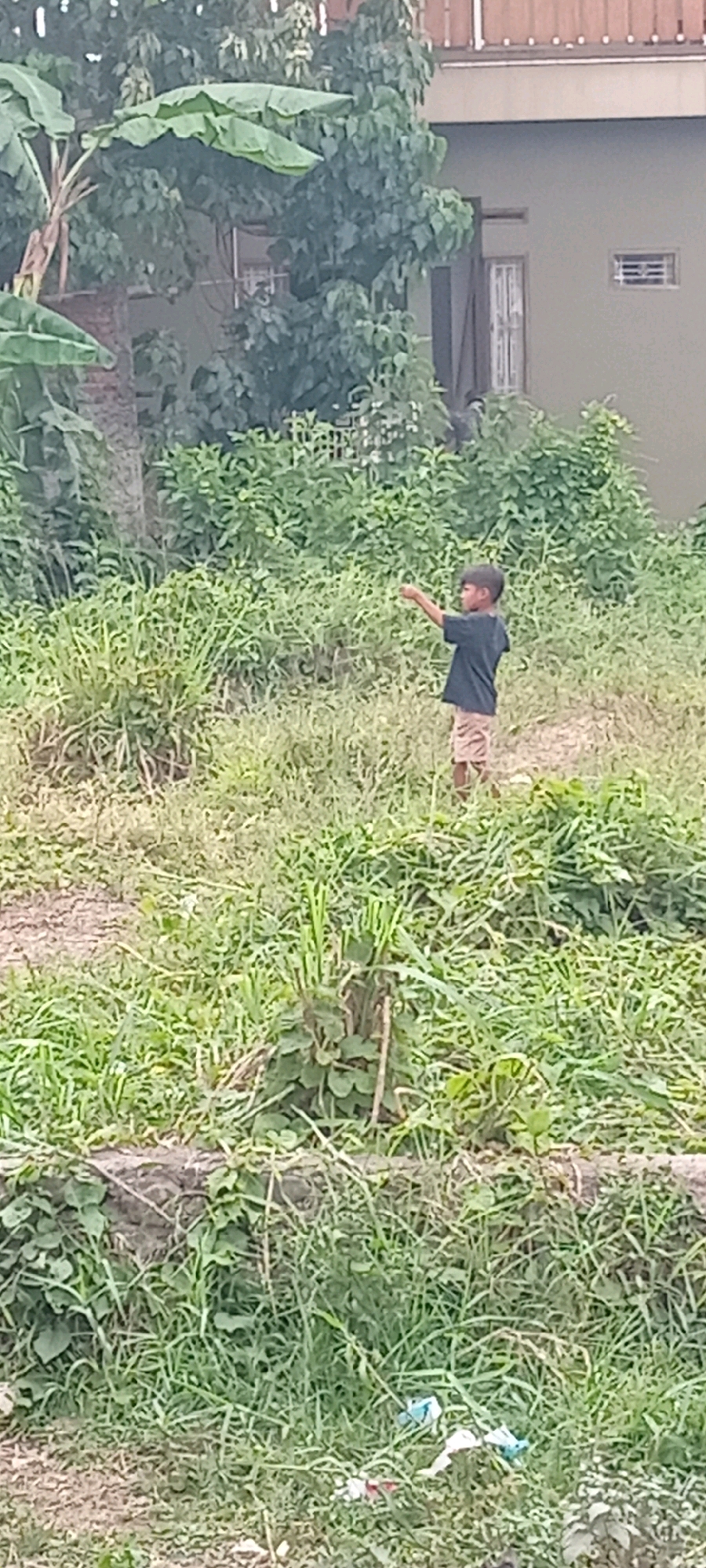 A young boy stands in overgrown grass, pointing to his left. He is wearing a dark t-shirt and patterned shorts. Behind him, a building with a light-colored wall and a wooden fence on top is visible, partially obscured by trees and lush vegetation. The scene appears to be an outdoor area in Tasikmalaya, Indonesia, characterized by dense greenery and a residential structure. The ground is covered with a mix of grass and dirt. A few pieces of trash are scattered in the lower right portion of the image.