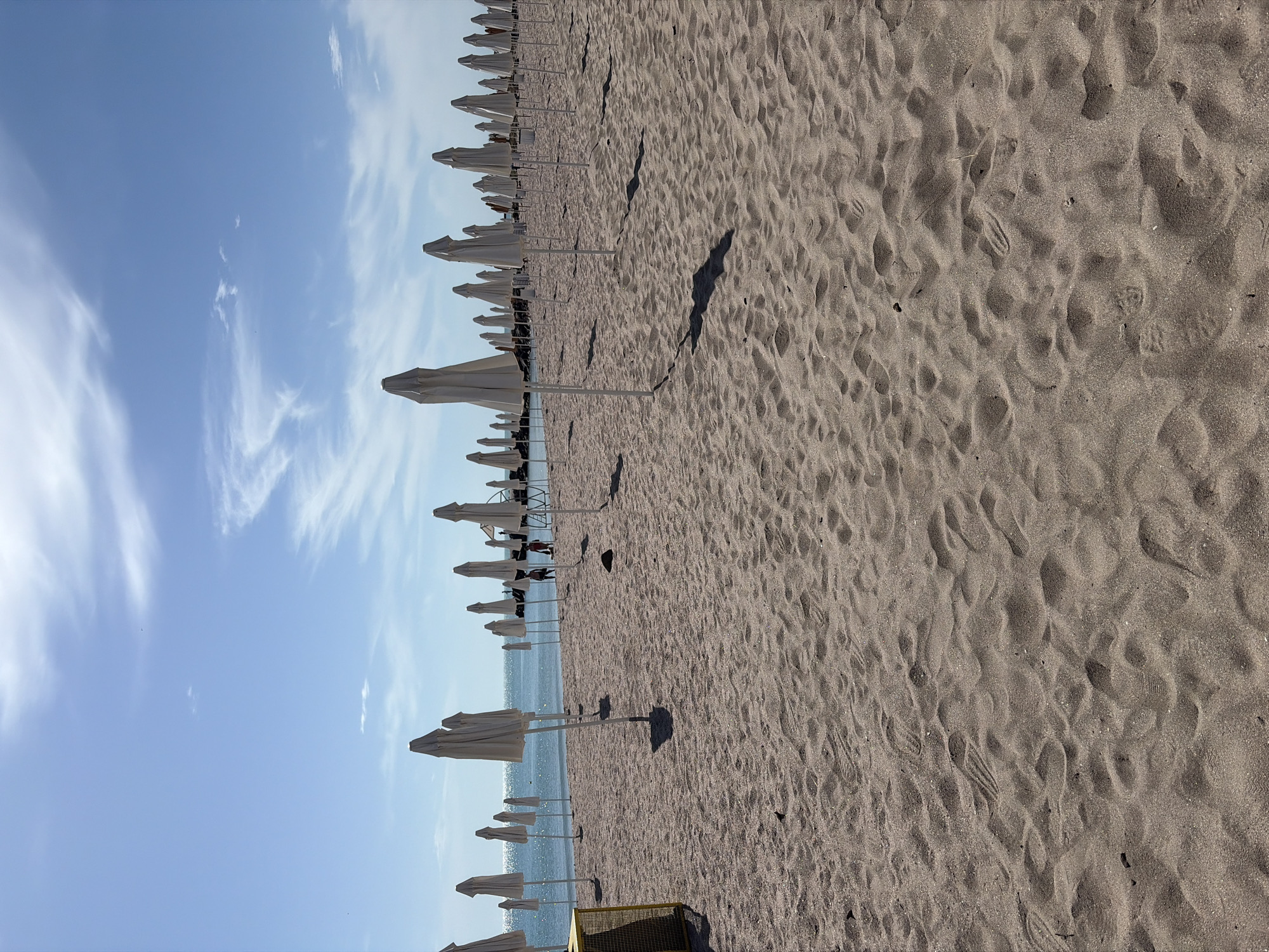 This image depicts a beach in Unknown, Unknown, filled with rows of white beach umbrellas. The umbrellas cast shadows on the sand. The sand itself is textured with footprints and wind patterns. The sky is light blue with wispy white clouds. The ocean is visible in the distance, appearing as a horizontal band of blue between the sand and the sky. There are no people clearly visible in the image.