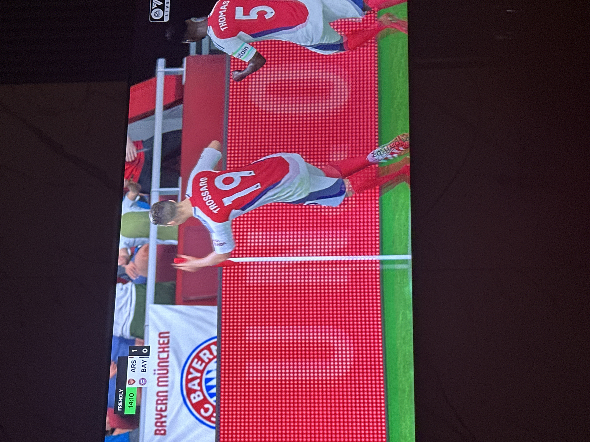 The image shows a screenshot from a soccer video game, displayed on a television screen. In the game, two players wearing red and white kits are visible on a field. One player, identified as "TROSSARD 19" on his jersey, is celebrating a goal, raising his arm. Another player, "THOMAS 5", is to his right, also in a celebratory pose. A score overlay in the top left corner shows "FRIENDLY 14:10 ARS 1 BAY 0". A banner with "BAYERN MÜNCHEN" and a club crest is visible in the background. The scene is illuminated by the bright display of the television. The image is taken from a slightly elevated angle, looking down at the screen. There is no discernible connection to Gwarinpa, Nigeria.