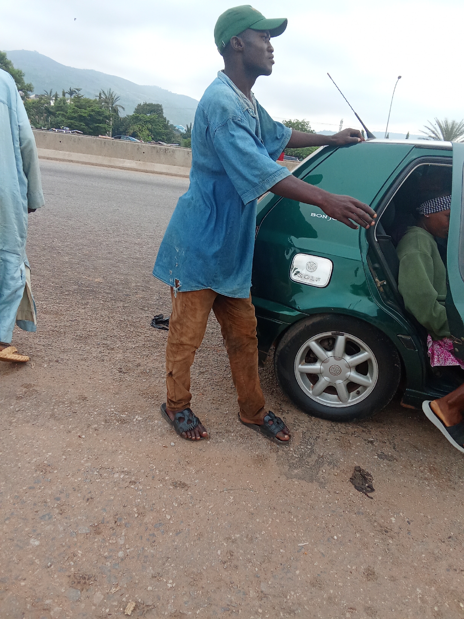 A young Black male wearing a blue denim shirt and brown pants, with a green baseball cap, stands next to a green Volkswagen Golf car on a dirt road in Kagini, Nigeria. The male is reaching towards the open passenger door. Inside the car, a person wearing a green sweatshirt and a patterned headscarf is visible. The car's "GOLF" emblem is clearly visible on the rear quarter panel. The background shows a mountainous landscape and trees. The ground is covered in gravel and dirt, with some dark debris scattered on the surface. Another person, partially visible on the left, is wearing a light blue robe.