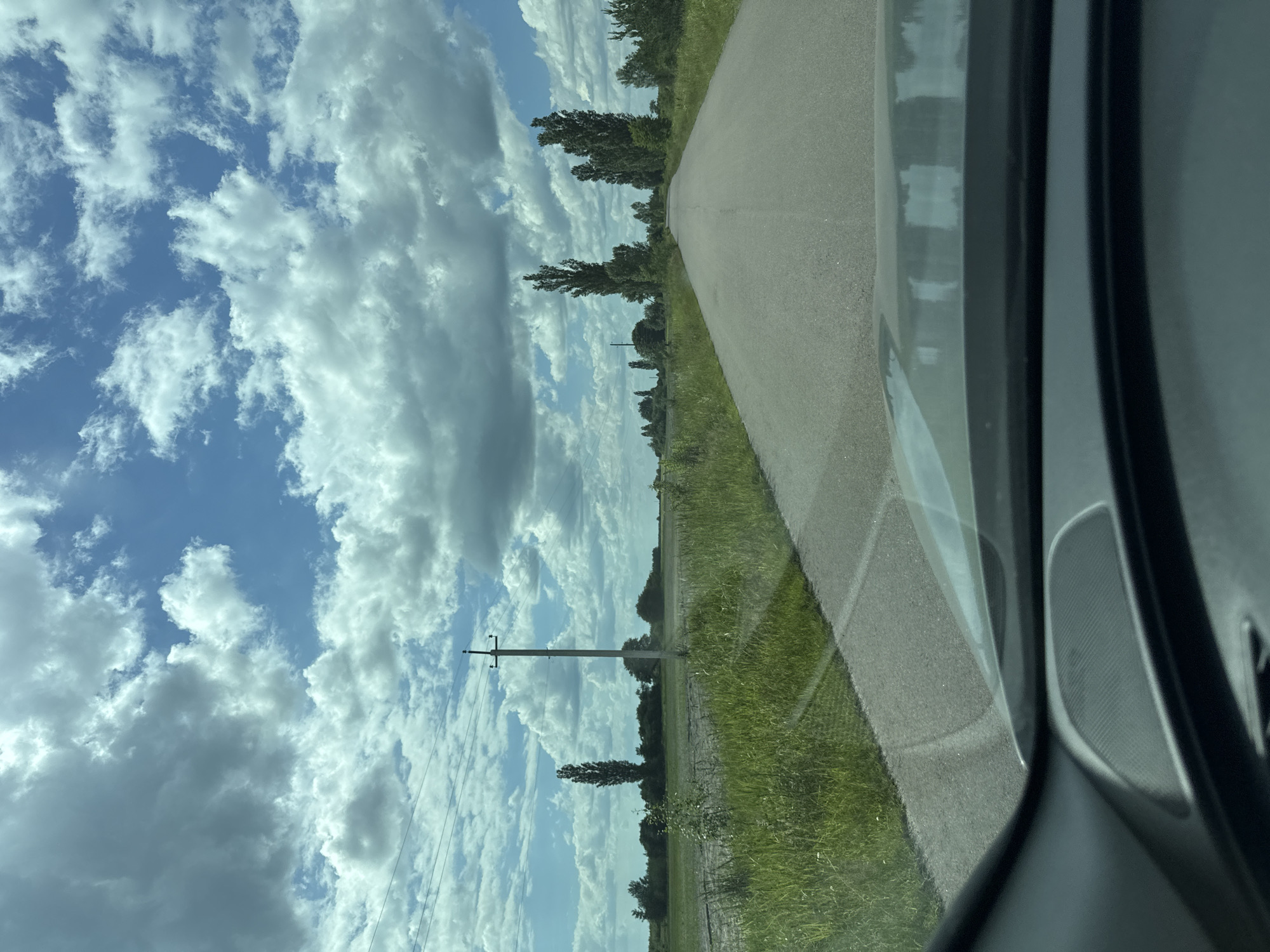 The image depicts a view from a vehicle traveling on a paved road through a rural landscape. The road stretches into the distance, flanked by grassy fields and scattered trees. Tall, slender trees, characteristic of poplars, are visible along the sides of the road and in the background. A single utility pole with power lines stands to the left of the road in the mid-ground. The sky is partly cloudy, with a mix of blue sky and white, puffy clouds. The foreground shows the dashboard and windshield of the vehicle, reflecting the sky and the road. The overall impression is one of a journey through a quiet, open countryside. The location is identified as Dovhe, Ukraine.
