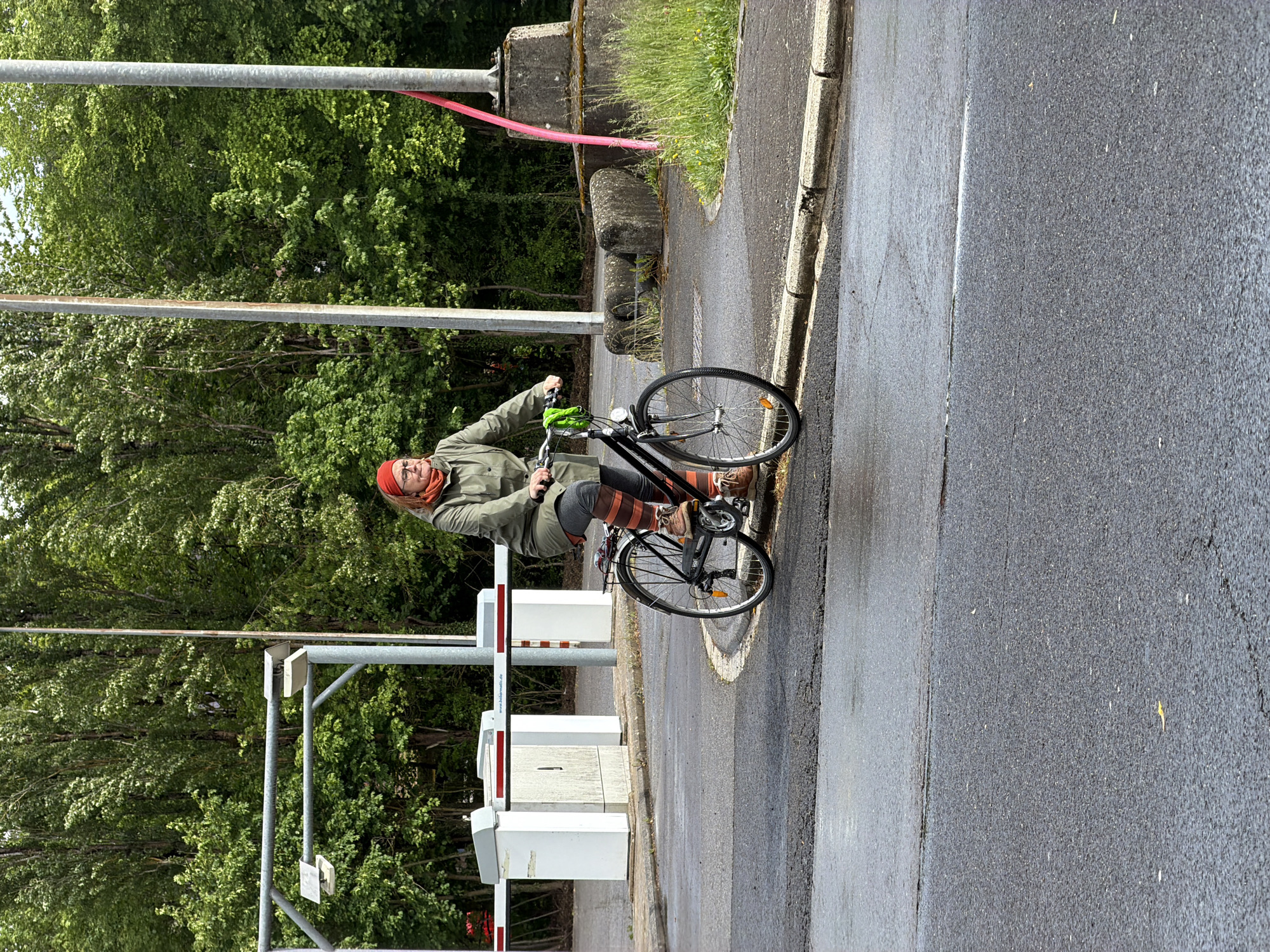 A middle-aged person on a black bicycle is stopped at a pedestrian crossing gate in Esch-sur-Alzette, Luxembourg. The person is wearing a red head covering, glasses, a green jacket, grey trousers, and brown and orange striped leg warmers. They also have brown shoes and green gloves. The crossing gate is composed of white and red barriers and is adjacent to a concrete barrier. Lush green trees are in the background. The road is wet and asphalt.