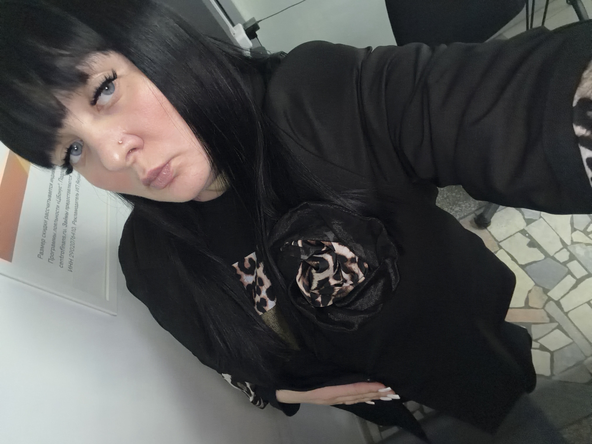 The image displays an adult female posing for a selfie inside what appears to be an office of Centrofinans, a microfinance organization, located in Chaikovsky, Russia.
The woman has dark, straight hair with bangs and blue eyes with prominent dark eyelashes. She features a small, light-colored stud piercing in her right nostril and her lips are slightly parted in a pout. She is wearing a long-sleeved black garment, which is accented with a large black fabric flower appliqué on her left chest. This appliqué incorporates leopard print fabric, a pattern also visible on a cuff or an underlayer on her left arm. Her left hand rests on her midsection, revealing long, almond-shaped fingernails painted white.
The background consists of a plain white wall, upon which a framed sign is mounted. The sign displays Russian text, explicitly mentioning "centrofinans.ru" and "Займы" (Loans), along with an INN (tax identification number), confirming the commercial nature and identity of the establishment. The floor is tiled with an irregular mosaic pattern of light grey, white, and beige geometric shapes. A dark, indistinct object, possibly part of a chair or furniture, is partially visible in the lower right corner. The lighting is consistent with an indoor artificial environment.