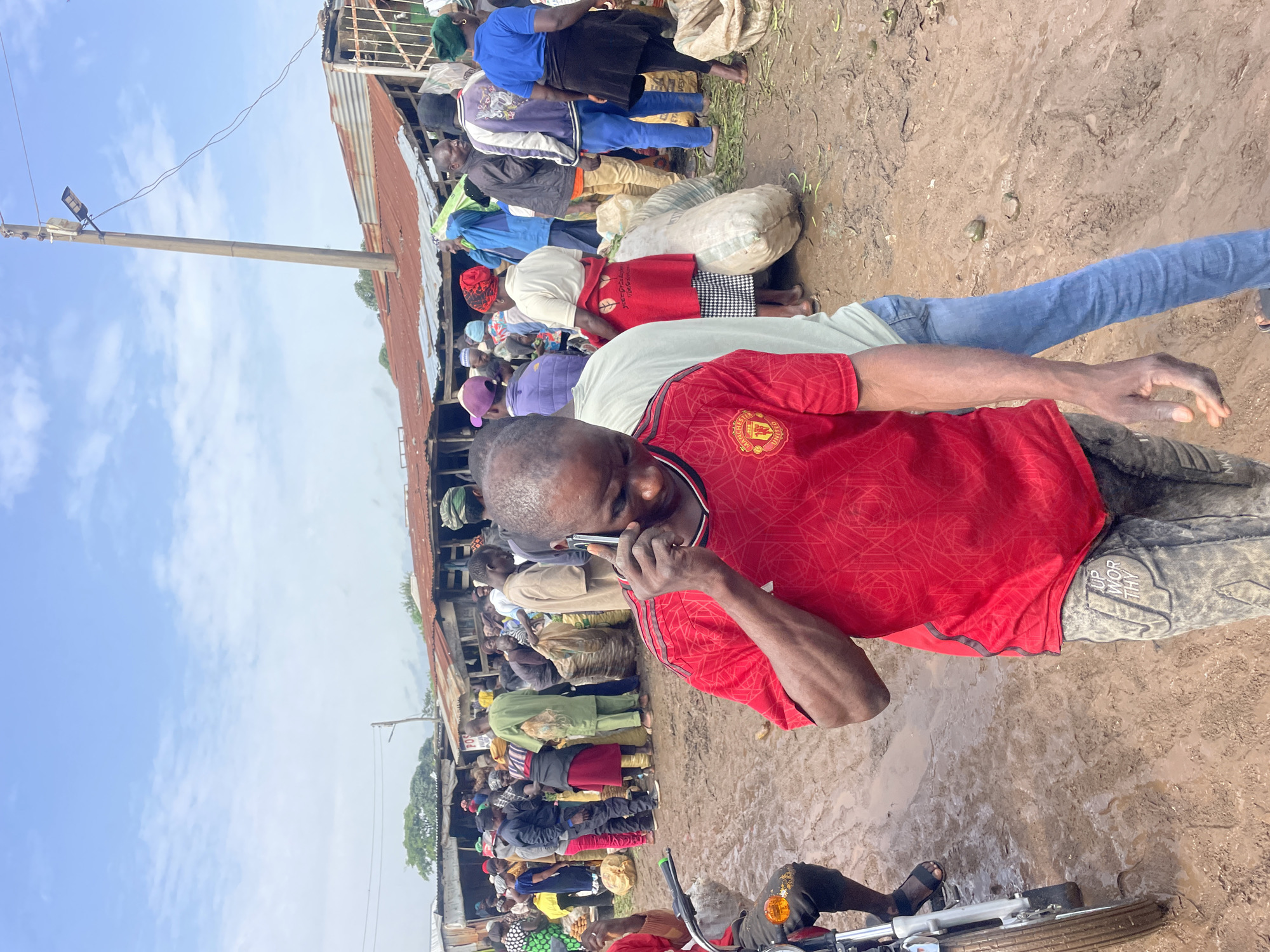 The image depicts a bustling outdoor market scene in Gura Top, Nigeria, under a cloudy sky. In the foreground, a man wearing a red Manchester United jersey and patterned pants is talking on a cell phone. His right arm is extended as he holds the phone to his ear, and his left hand is visible. Behind him, the market is filled with numerous people engaged in various activities. Several individuals are carrying or standing near large sacks, suggesting the sale or transport of goods. The ground is muddy, indicating recent rain or a naturally damp environment. In the background, market stalls with corrugated metal roofs line the area. A utility pole with a solar-powered light fixture is prominently visible. The overall atmosphere is one of active commerce and daily life in a Nigerian market.
