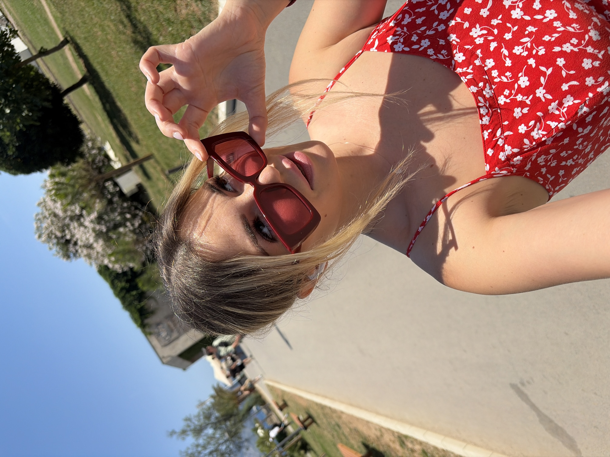 The image shows a young woman with blonde hair wearing red-framed sunglasses and a red floral sundress. She is holding her sunglasses up with her right hand. The woman is outdoors, with a paved walkway and grassy areas visible in the background. There are trees, a building, and a body of water in the distance. People are walking on the walkway in the background. The scene appears to be in Budva, Montenegro, likely on a sunny day.
